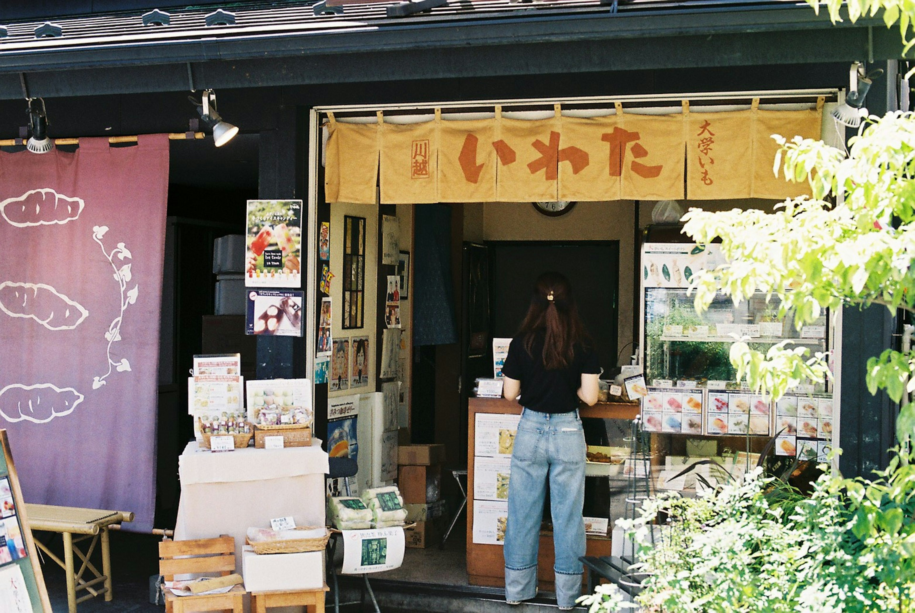 小さな店舗の外観で、木製の看板に「いわた」と書かれている。店の前には商品が並び、女性が作業をしている。