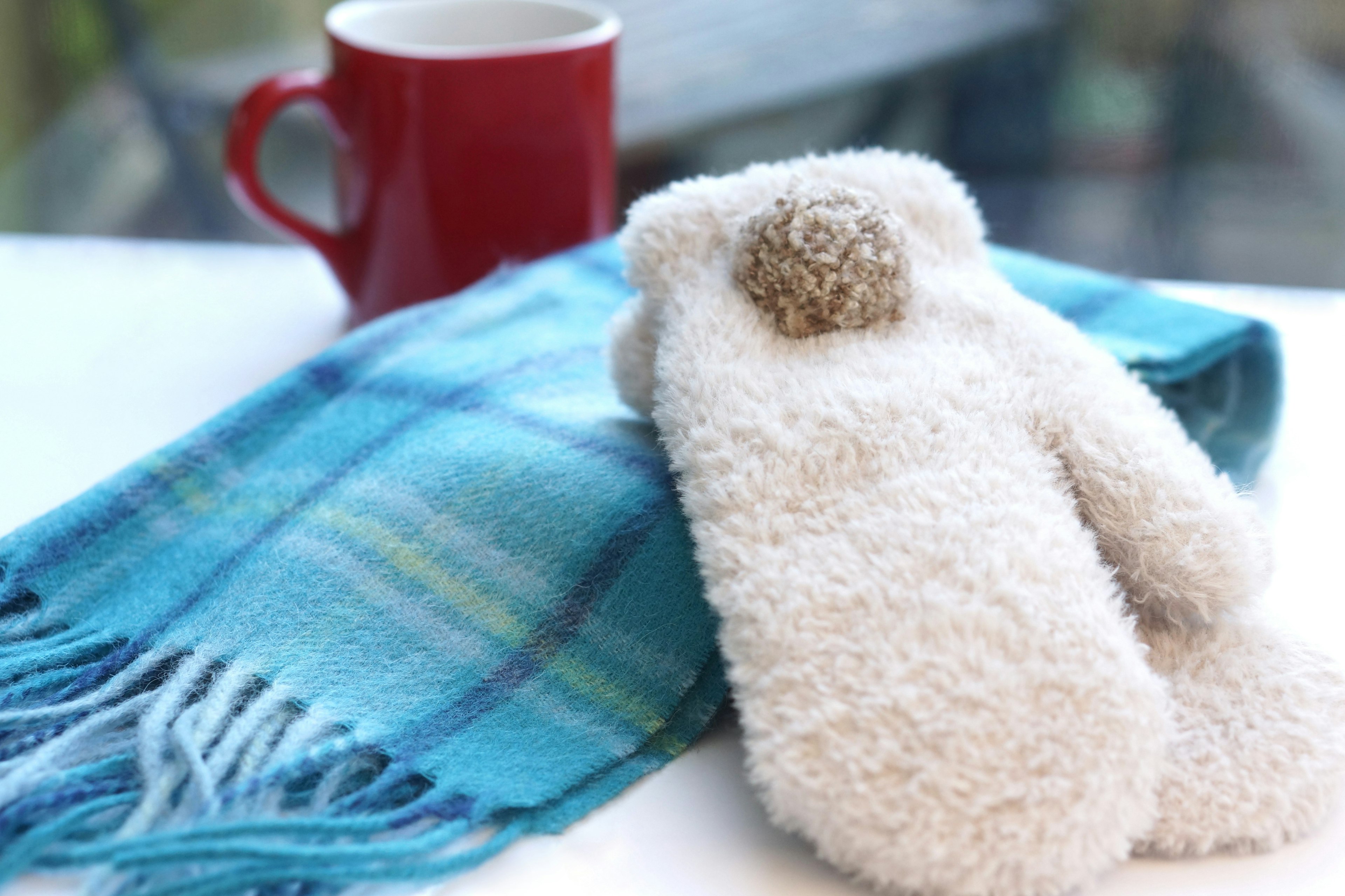 White mittens beside a red mug and a blue checkered scarf