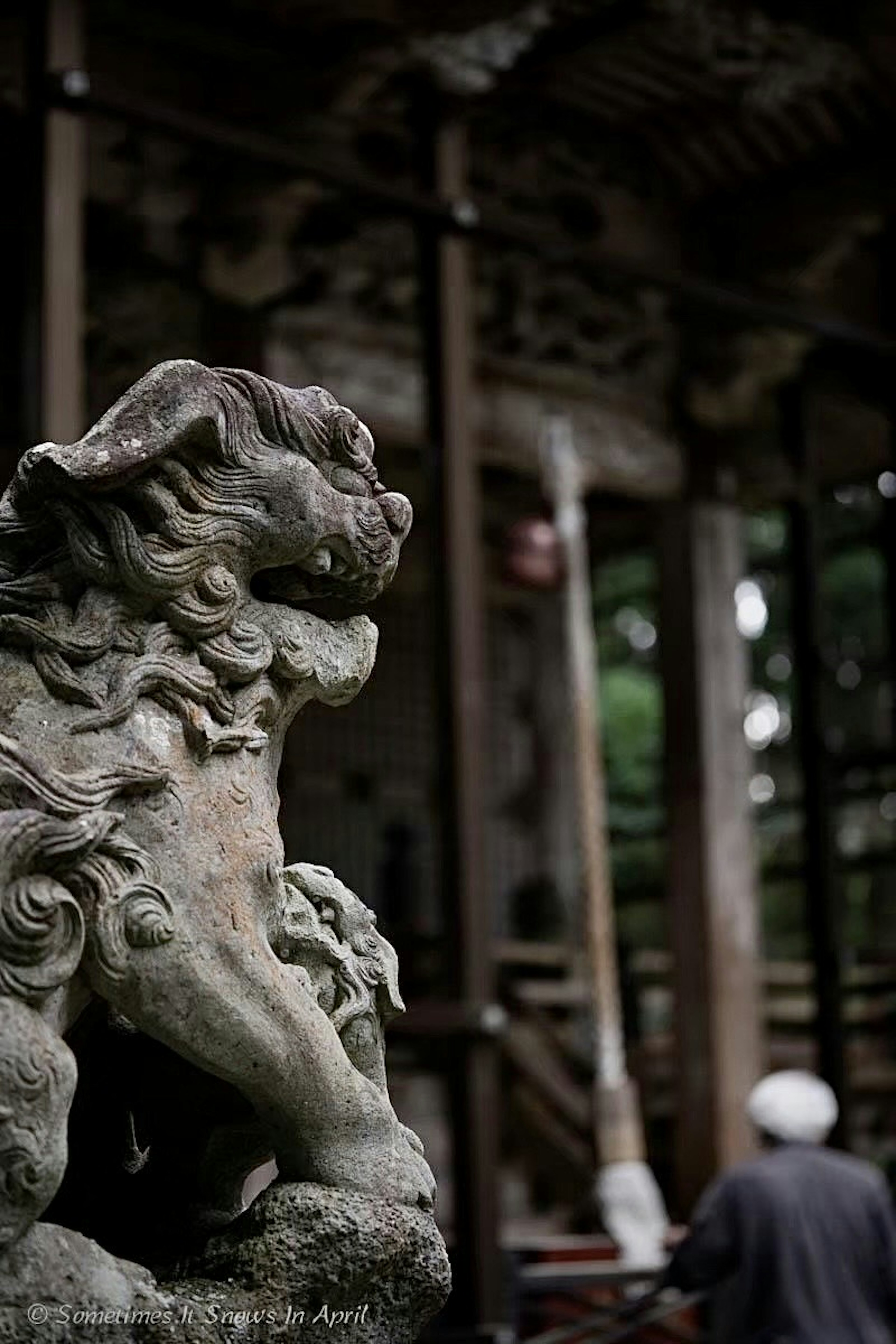 Una statua di leone guardiano in pietra davanti a un santuario con una figura sfocata di una persona sullo sfondo