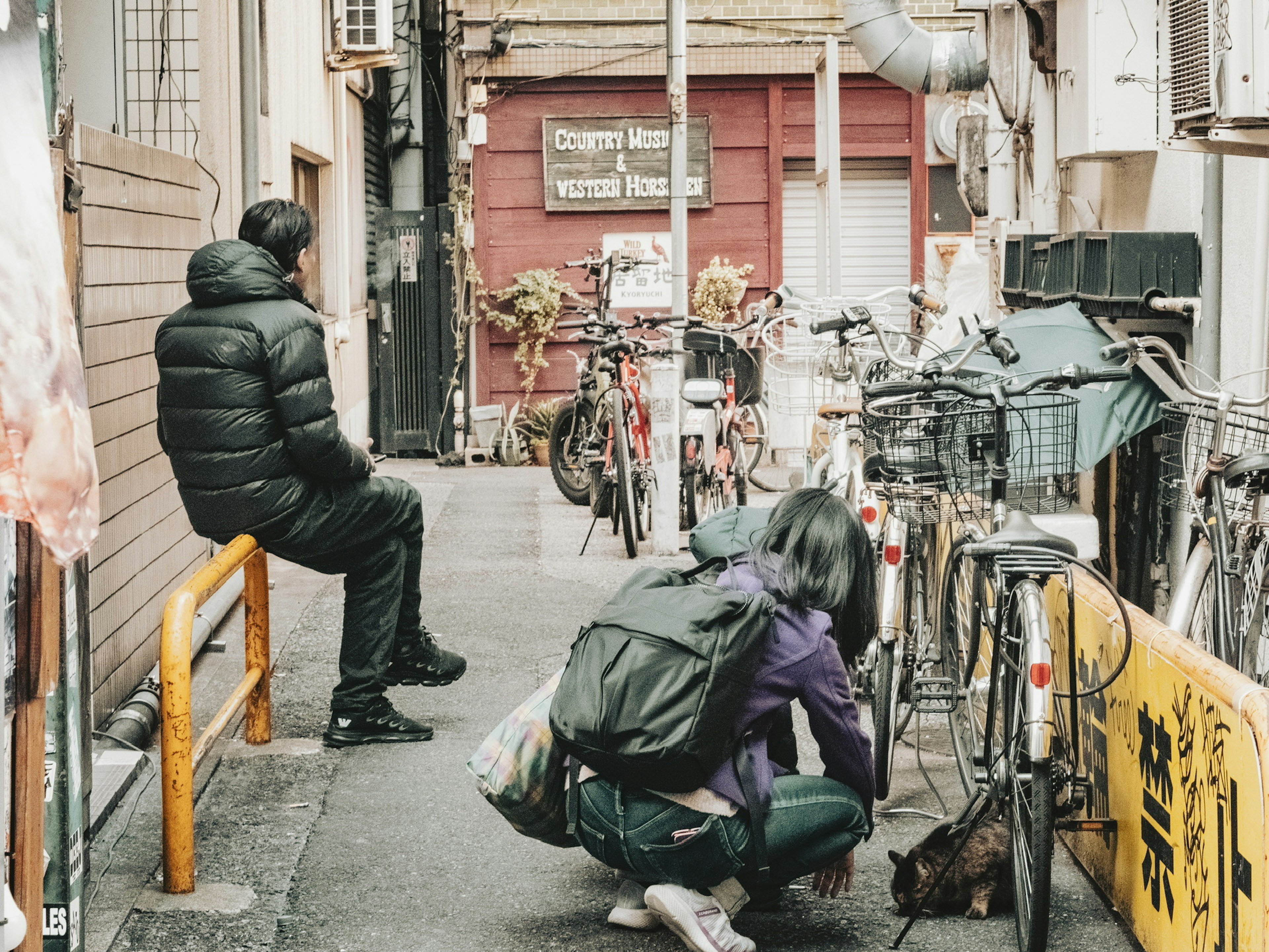 街の狭い通りに座っている男性と、地面にしゃがんでいる女性が自転車の近くにいる風景