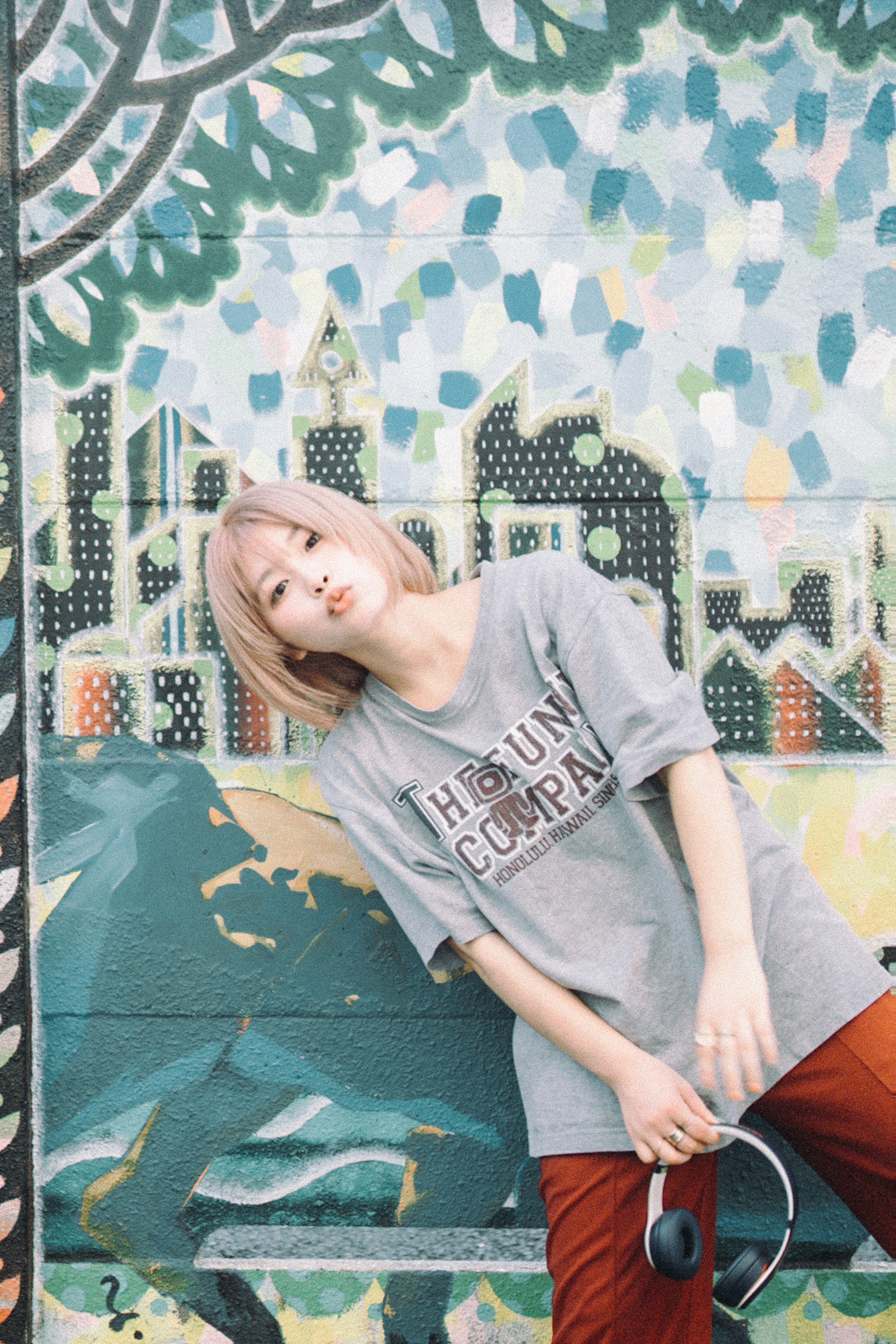 Joven mujer con camiseta gris y pantalones naranjas posando frente a un mural colorido