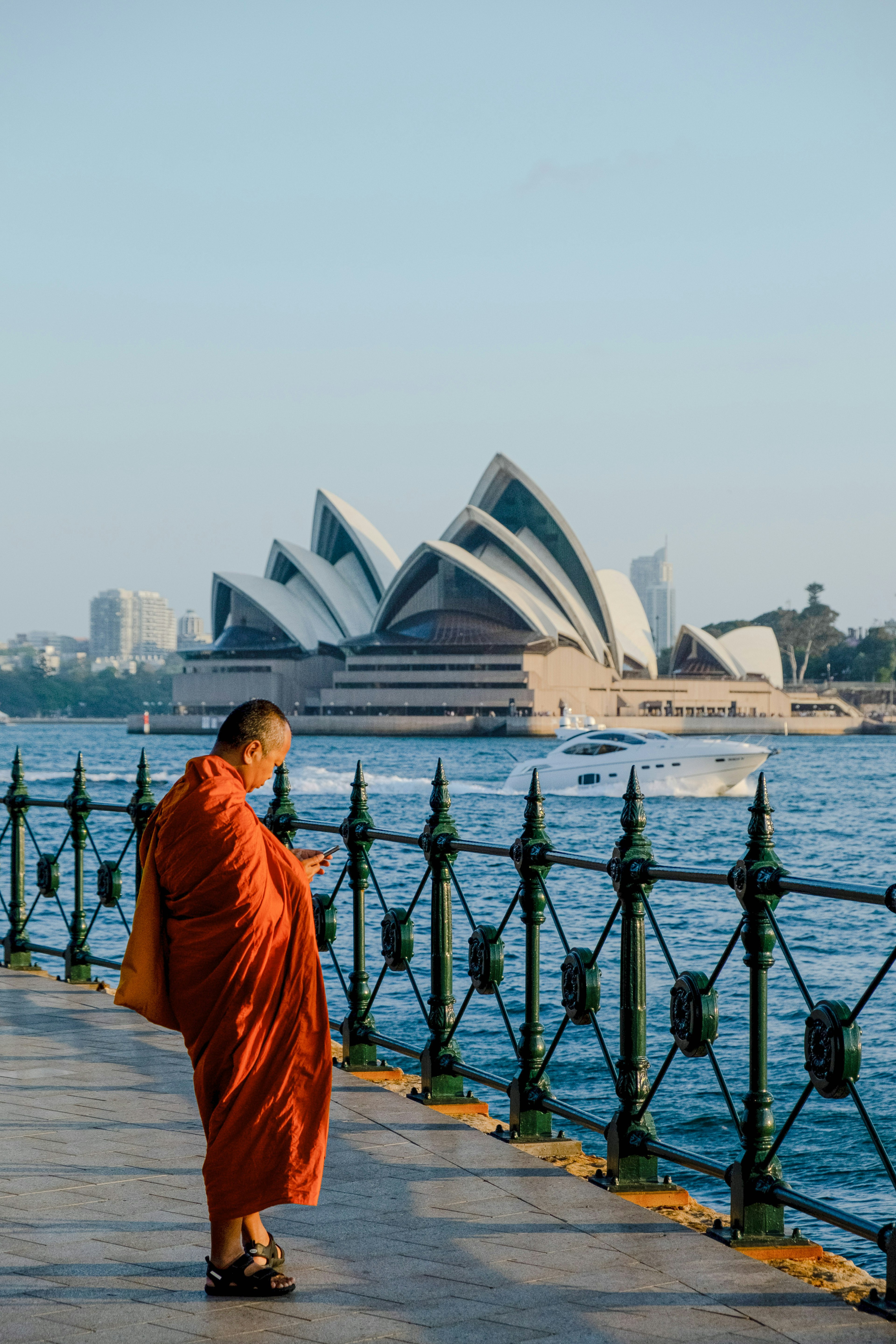 Seorang biksu dalam jubah oranye berjalan di tepi air dengan Sydney Opera House di latar belakang