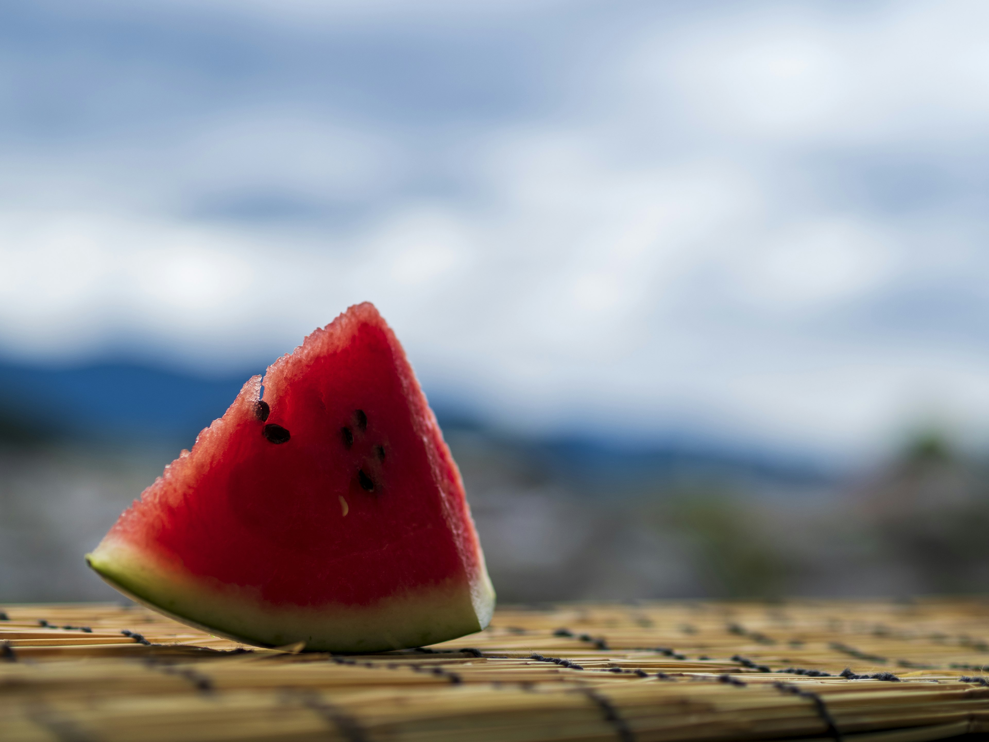 A slice of watermelon on a wooden table with a blurred blue sky and mountain silhouette in the background