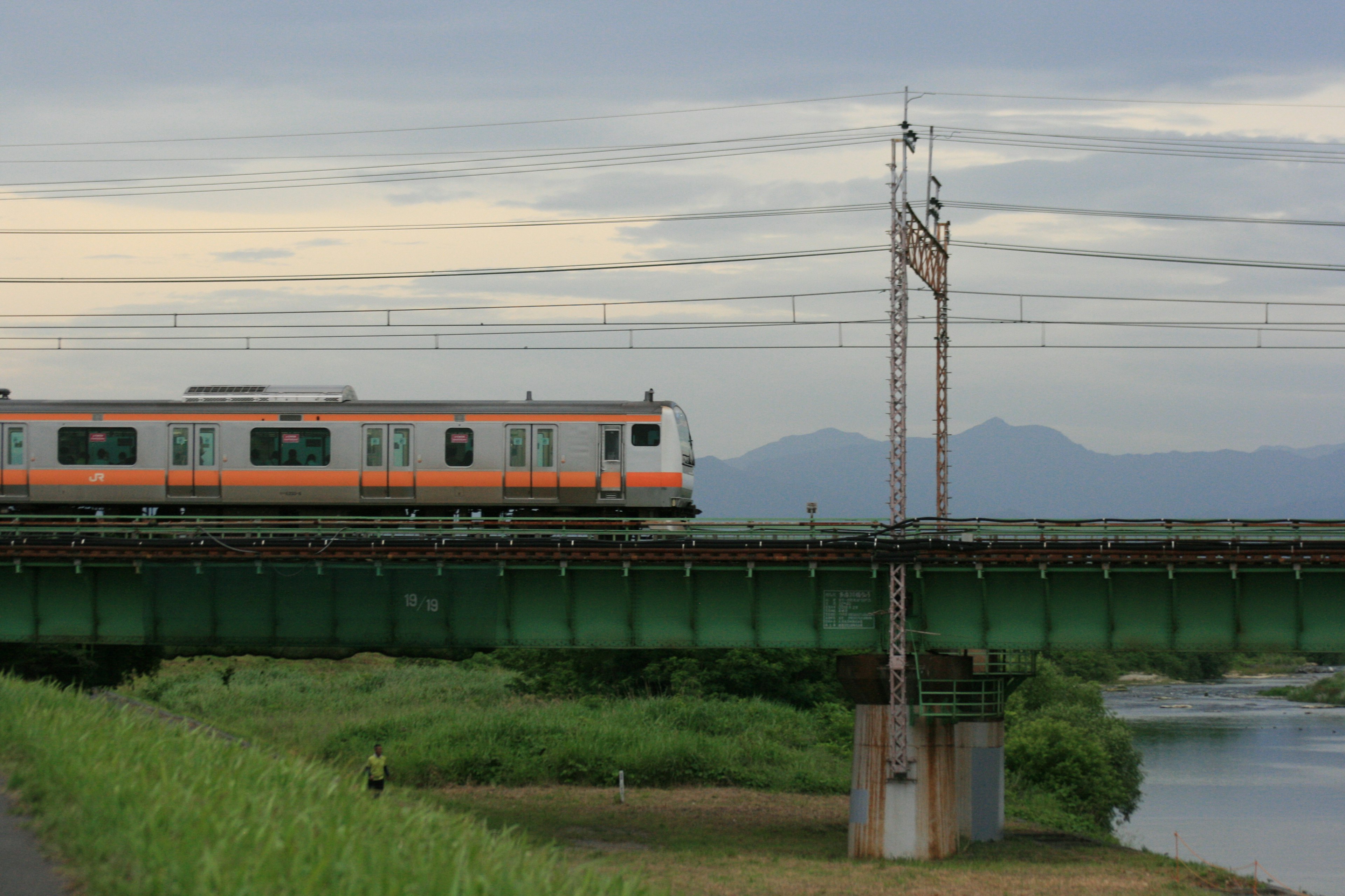 An orange train crossing a green bridge over a river with mountains in the background