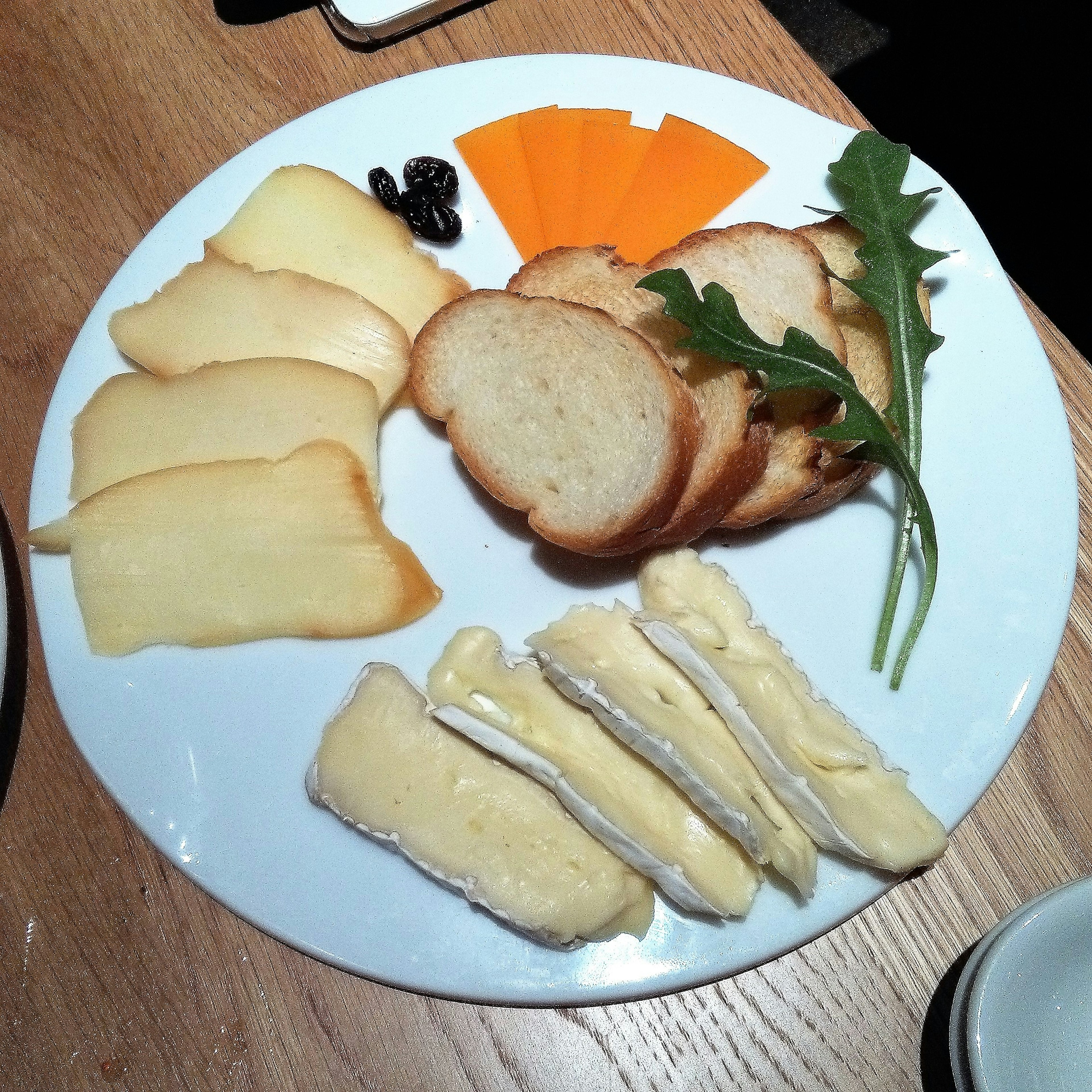 Plate featuring various cheeses, slices of bread, carrot strips, arugula, and dried fruit