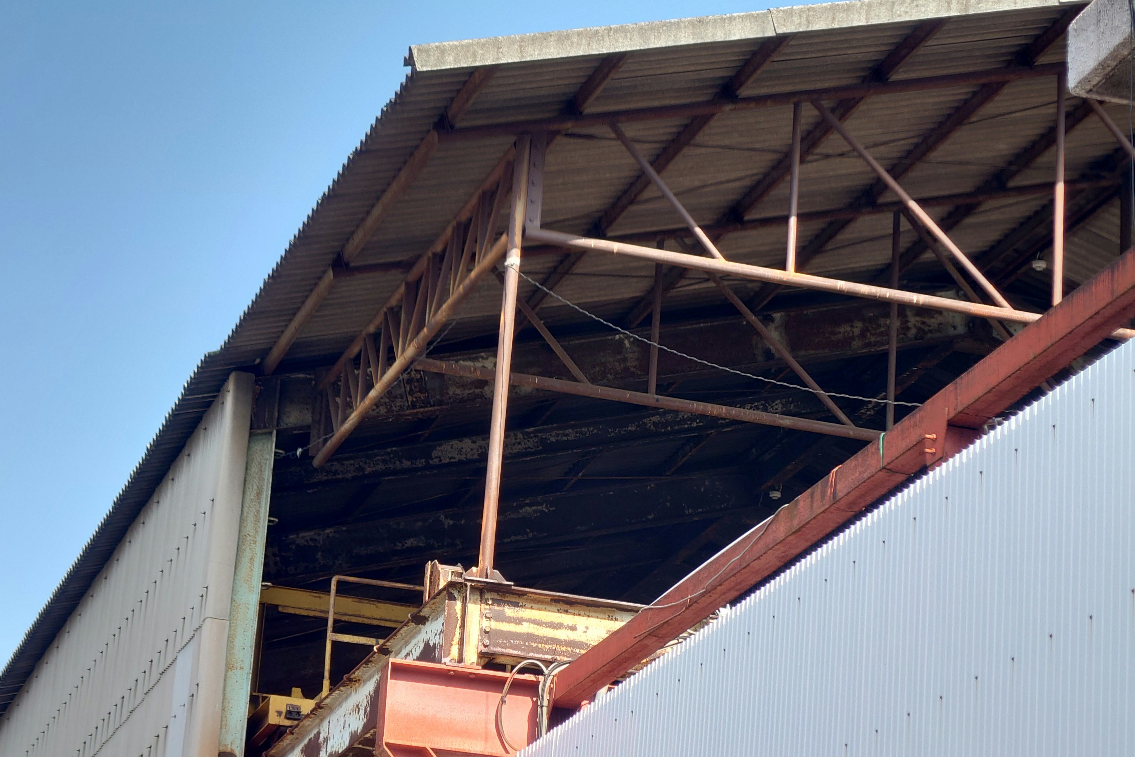 Photo d'une structure de toit d'usine montrant des poutres métalliques et des matériaux anciens sous un ciel bleu