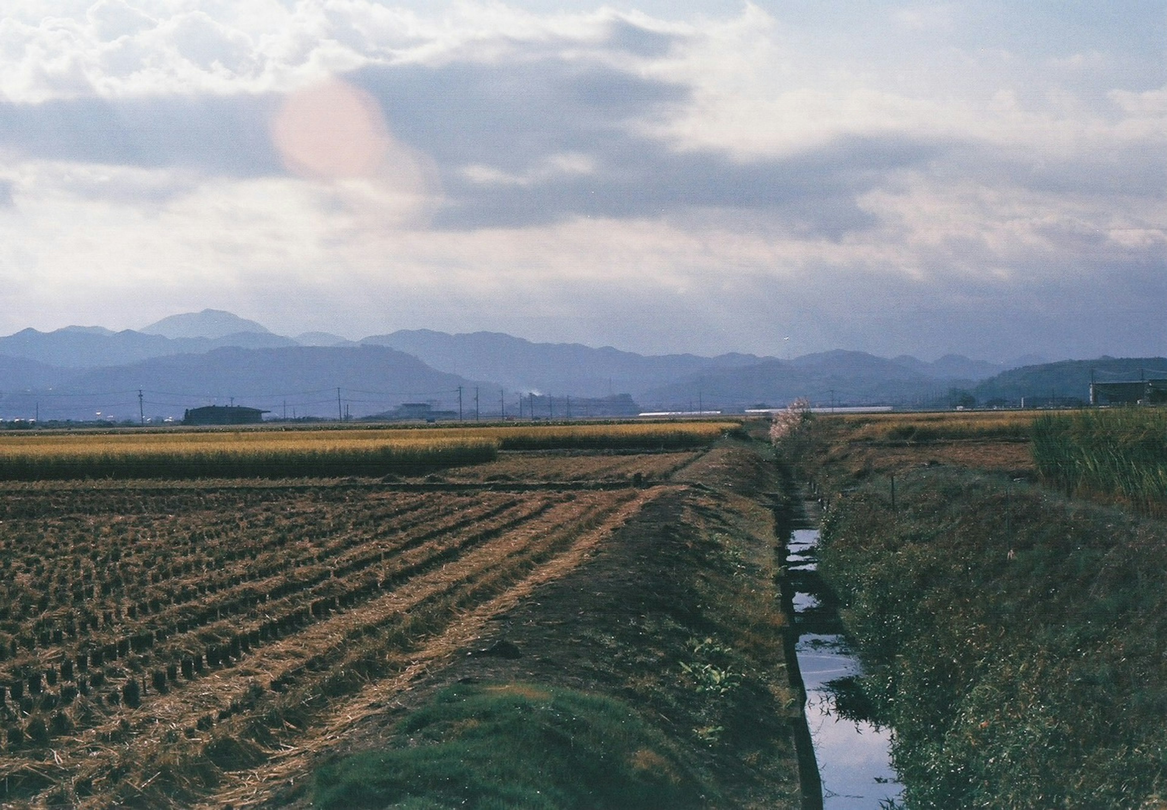 Paysage de rizières et d'un ruisseau sous un ciel nuageux avec des montagnes en arrière-plan