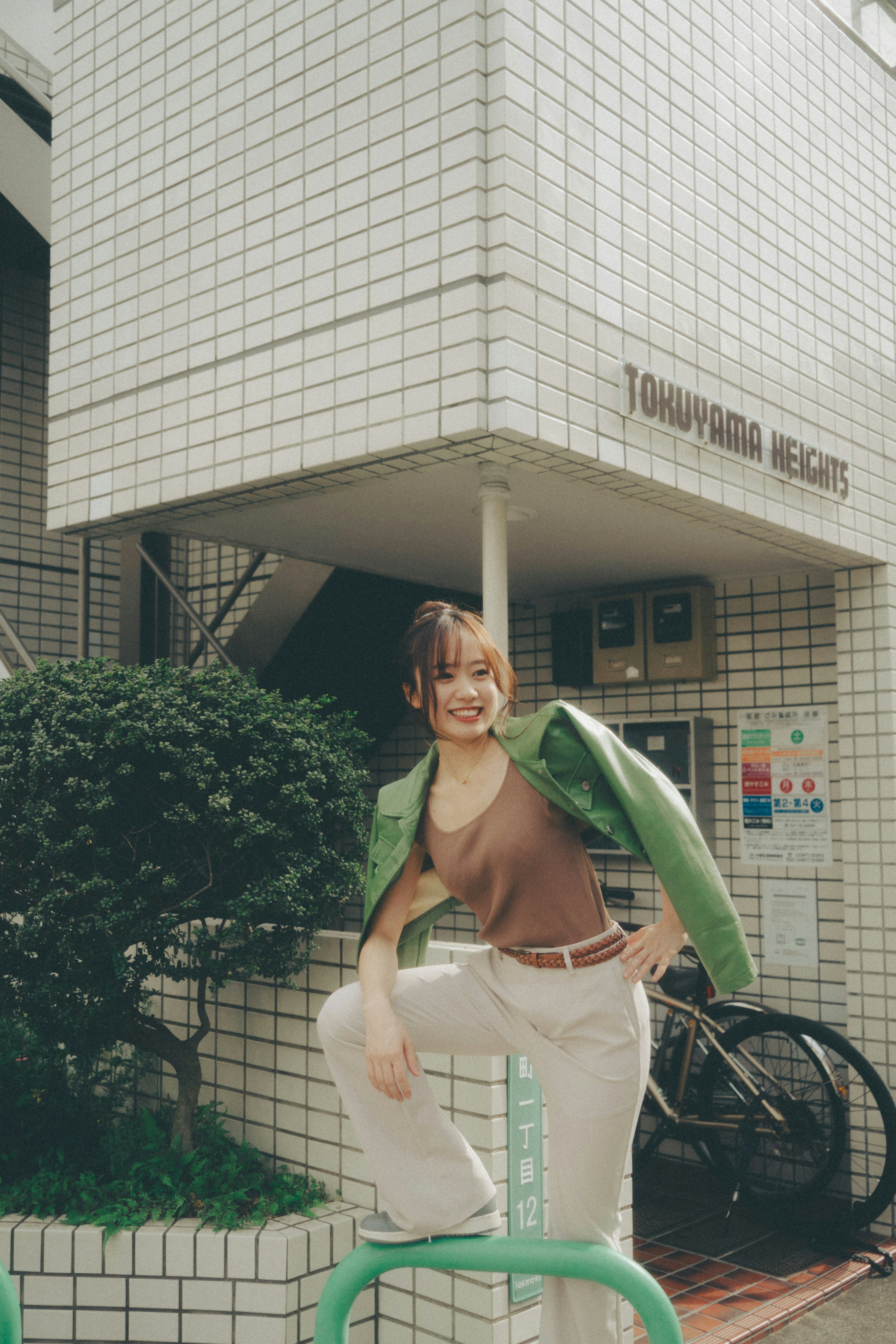 Femme en veste verte posant devant un bâtiment carrelé avec de la verdure