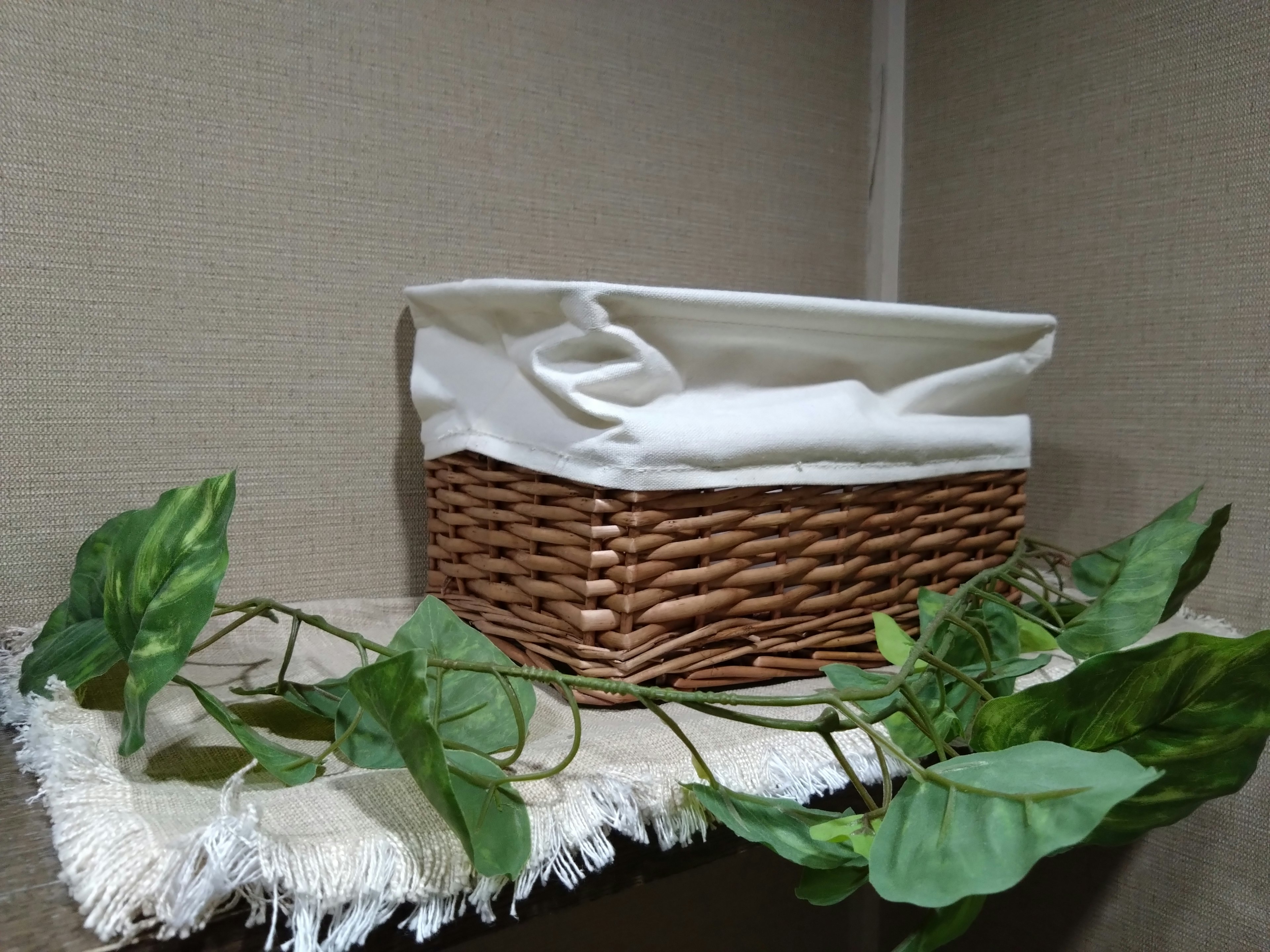 A wicker basket with a white fabric cover placed on a shelf with green plants nearby