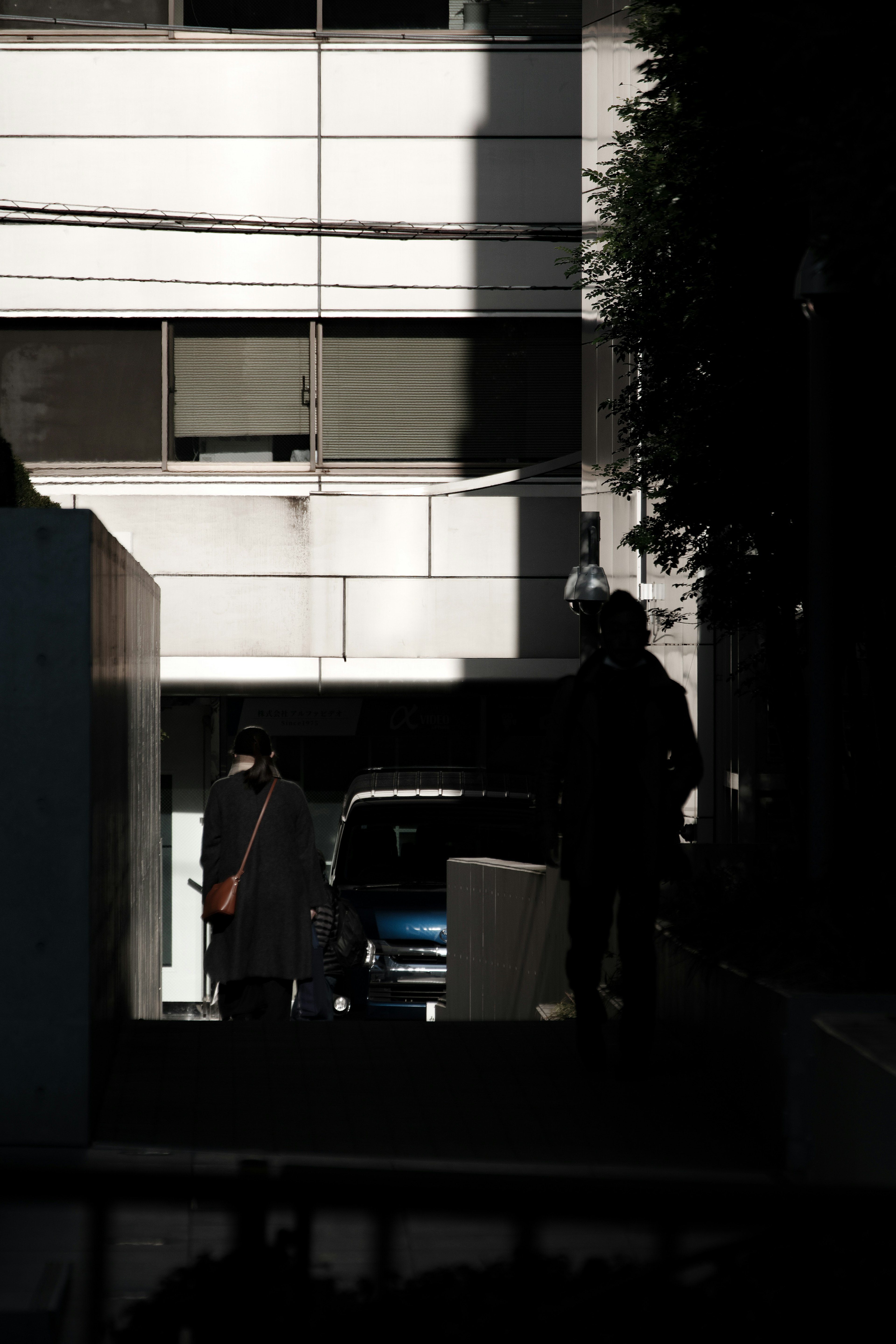 Siluetas de personas y un coche azul en la sombra de un edificio blanco