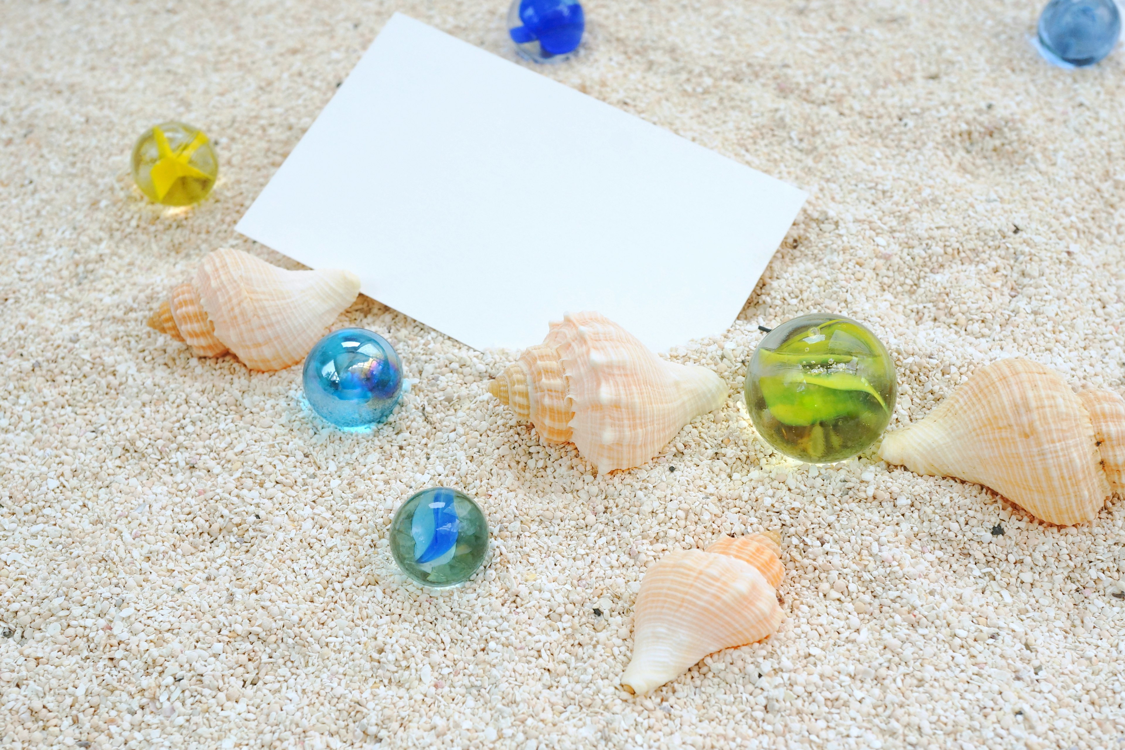 A blank card placed among seashells and colorful marbles on sandy beach