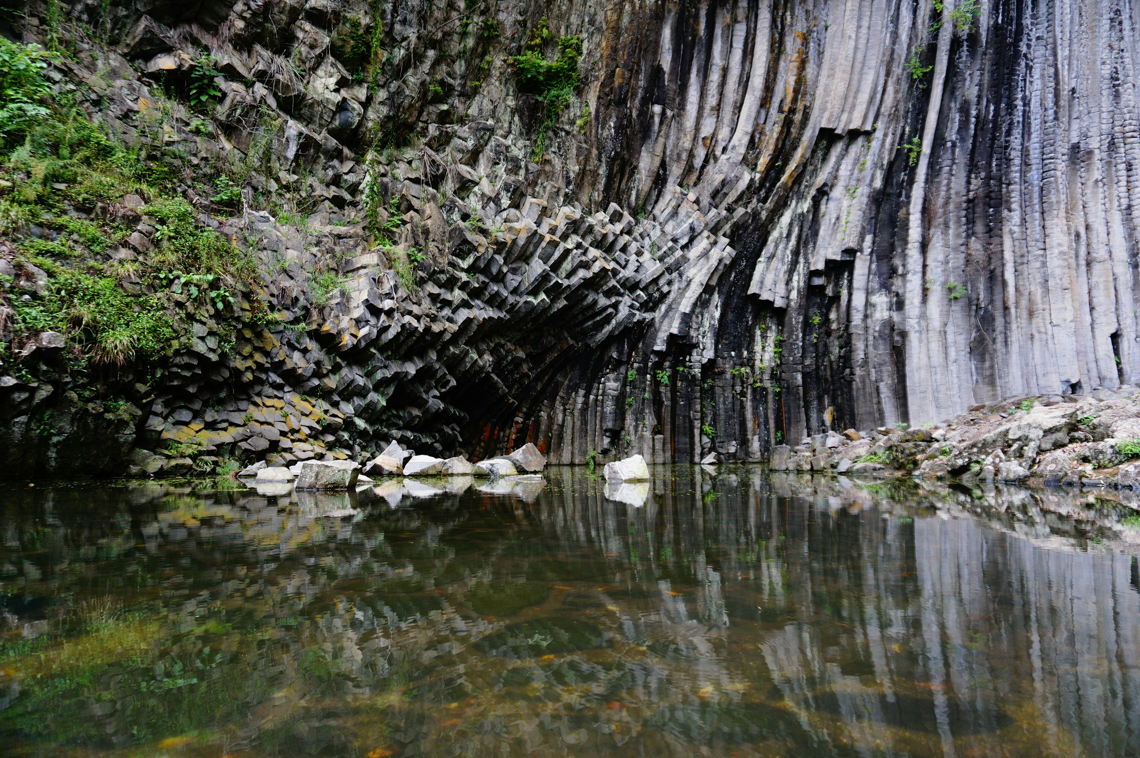 Columnar jointed rock formation reflected in calm water with lush greenery