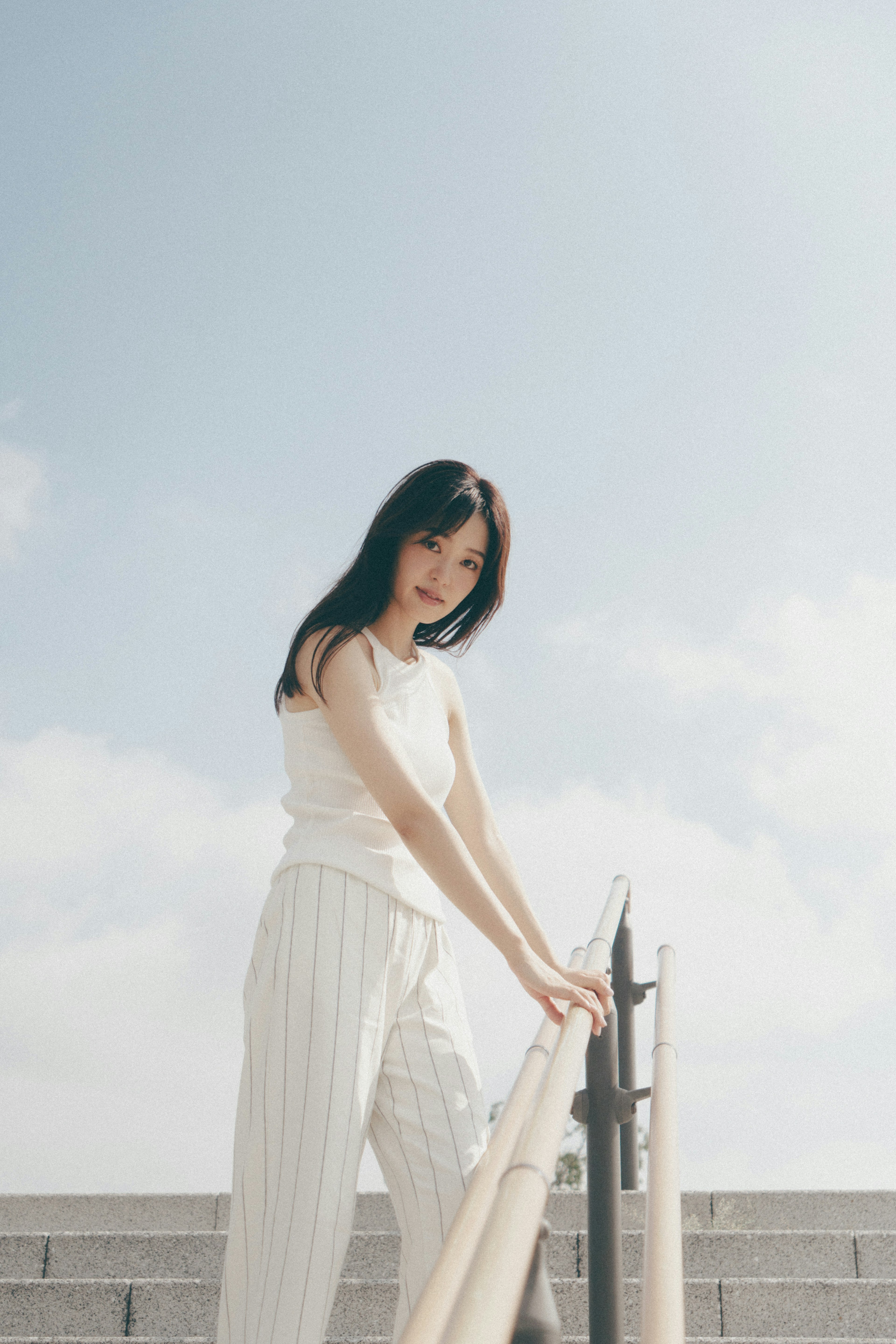 Woman in a white tank top leaning on a railing under a bright sky