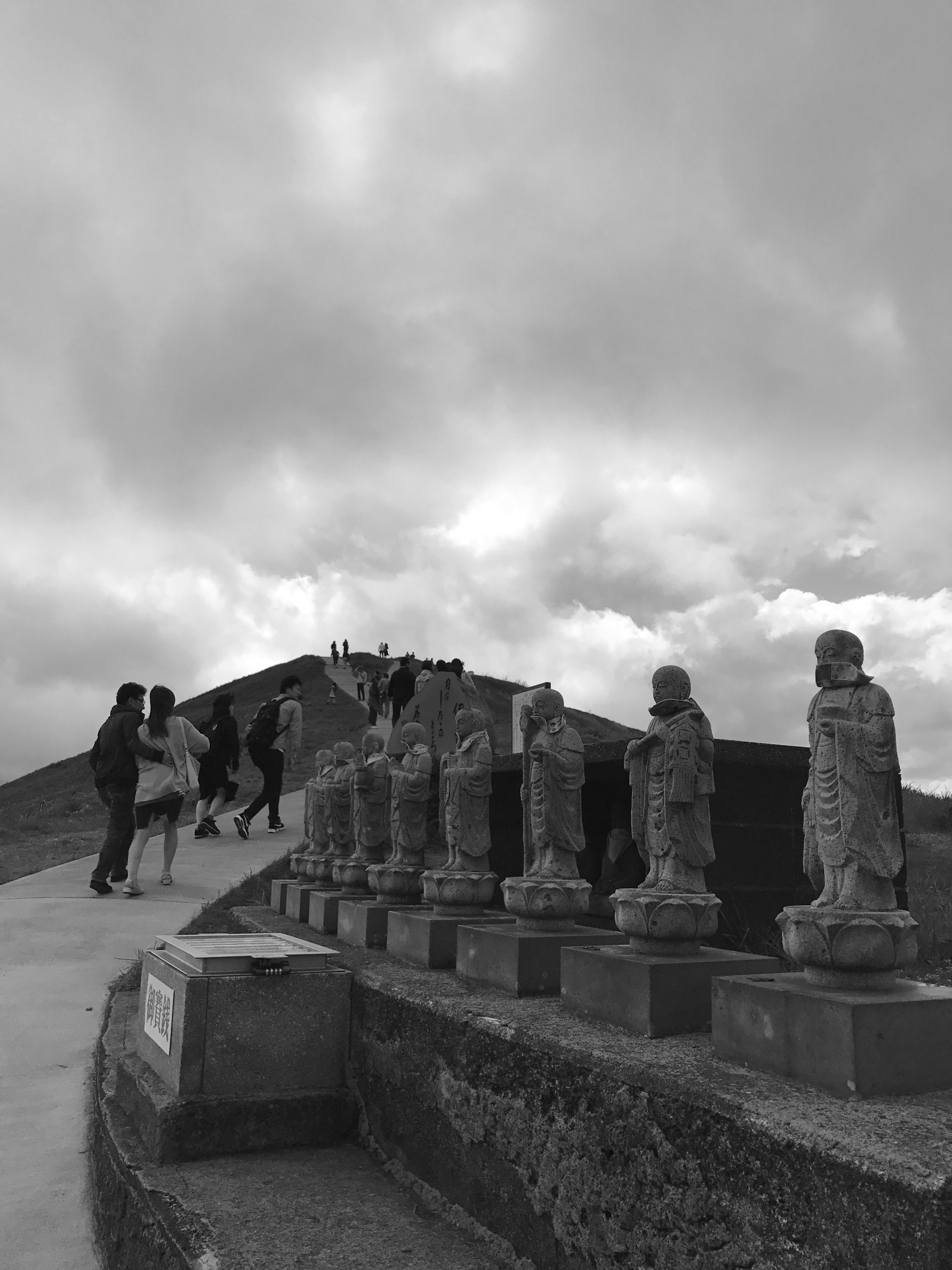 A scene with stone statues lined up under a cloudy sky with people walking