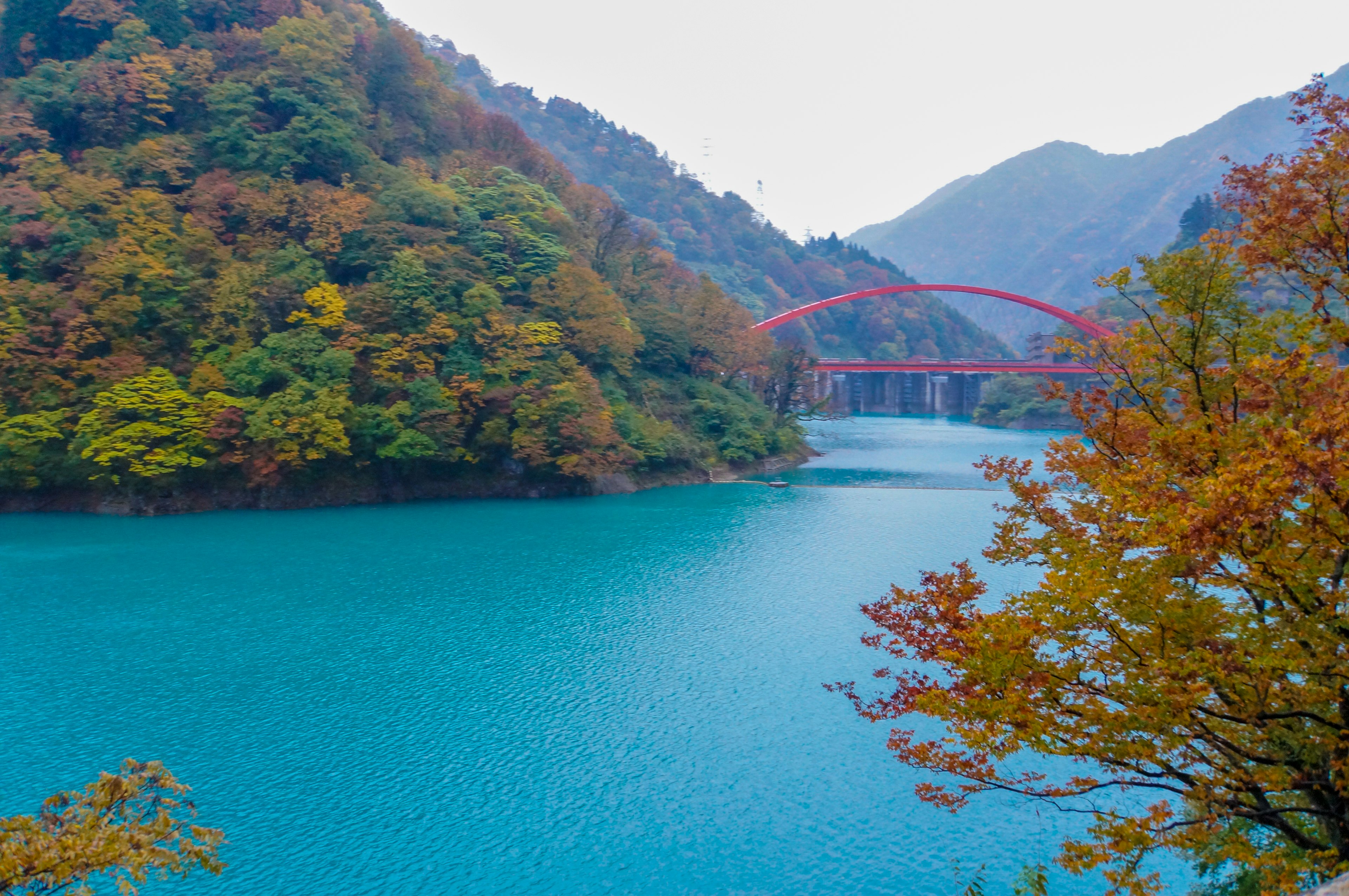 Malersicher Blick auf einen türkisfarbenen See umgeben von Herbstlaub und einer roten Brücke