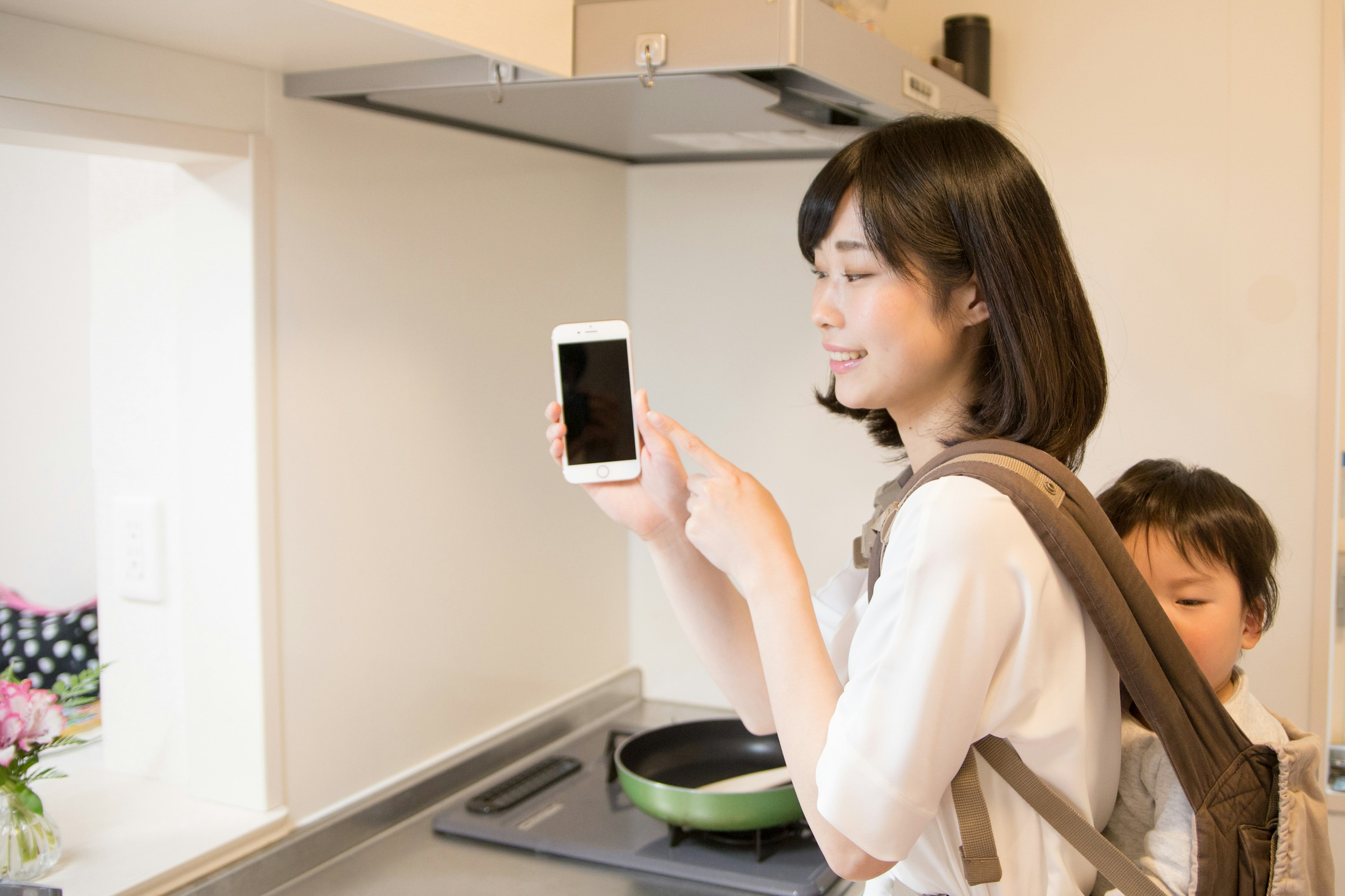 A woman holding a smartphone in a kitchen with a child behind her