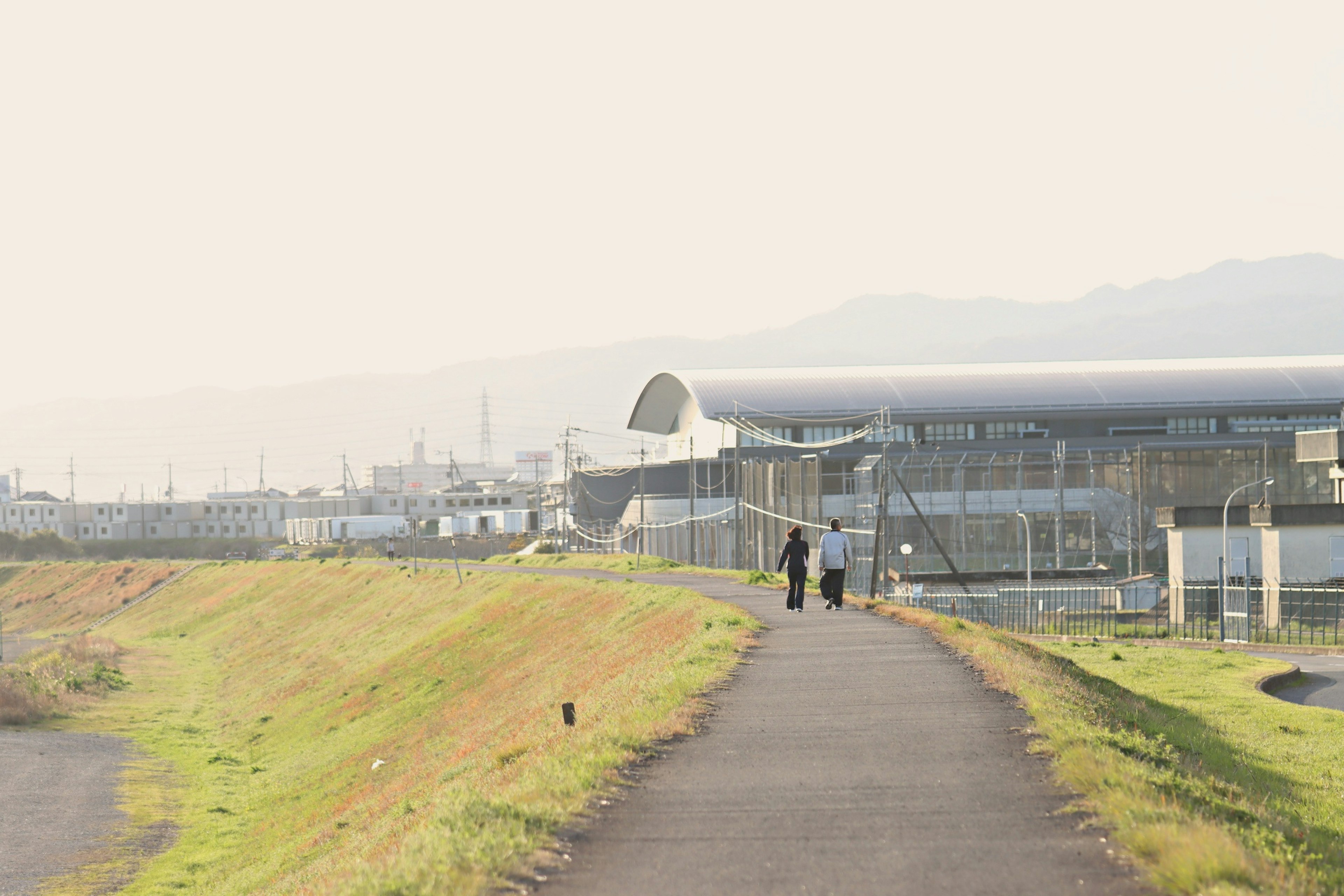 川沿いの道を歩く二人の人物と近くの建物が見える風景