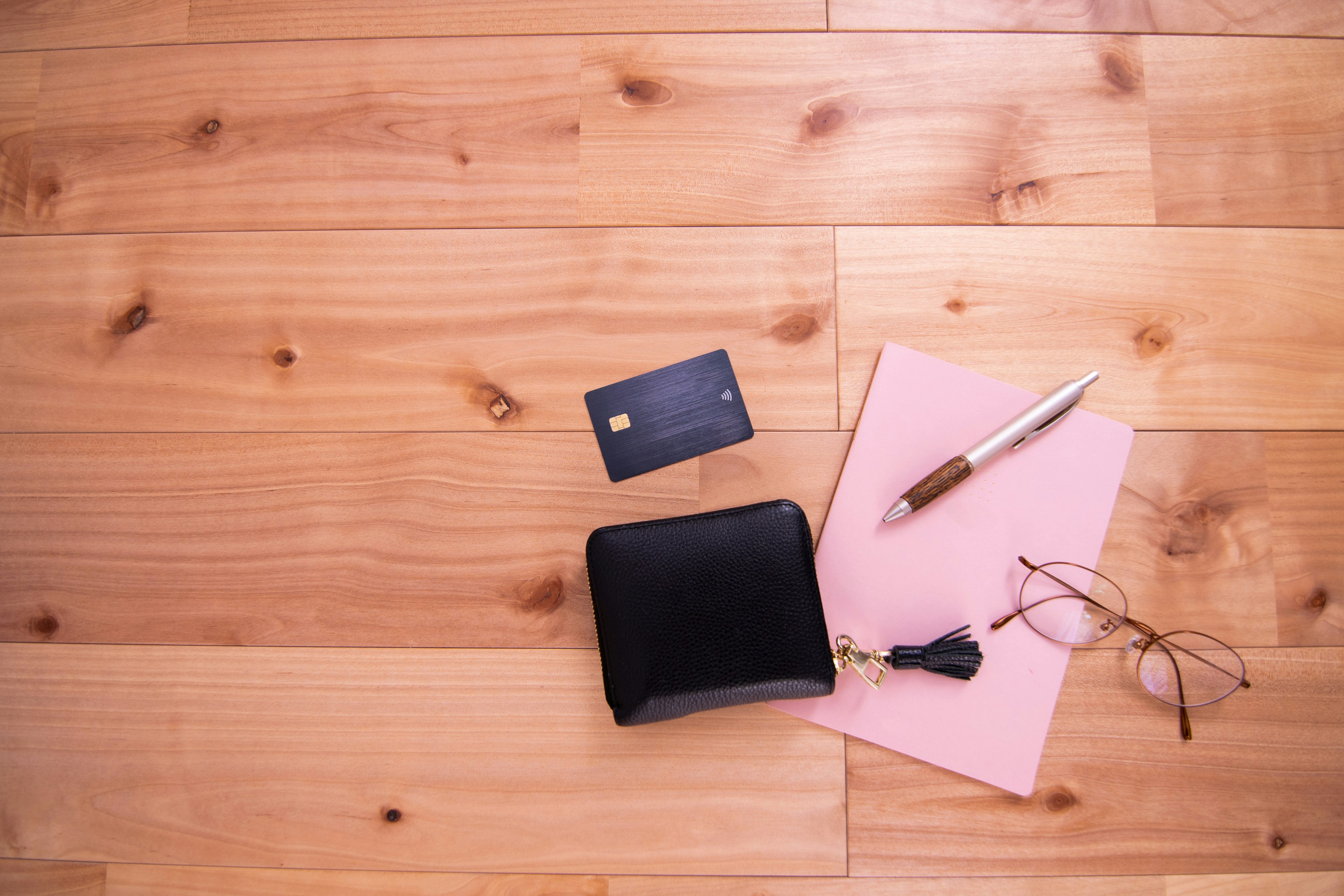 A black wallet with a credit card pink notebook pen and glasses arranged on a wooden floor