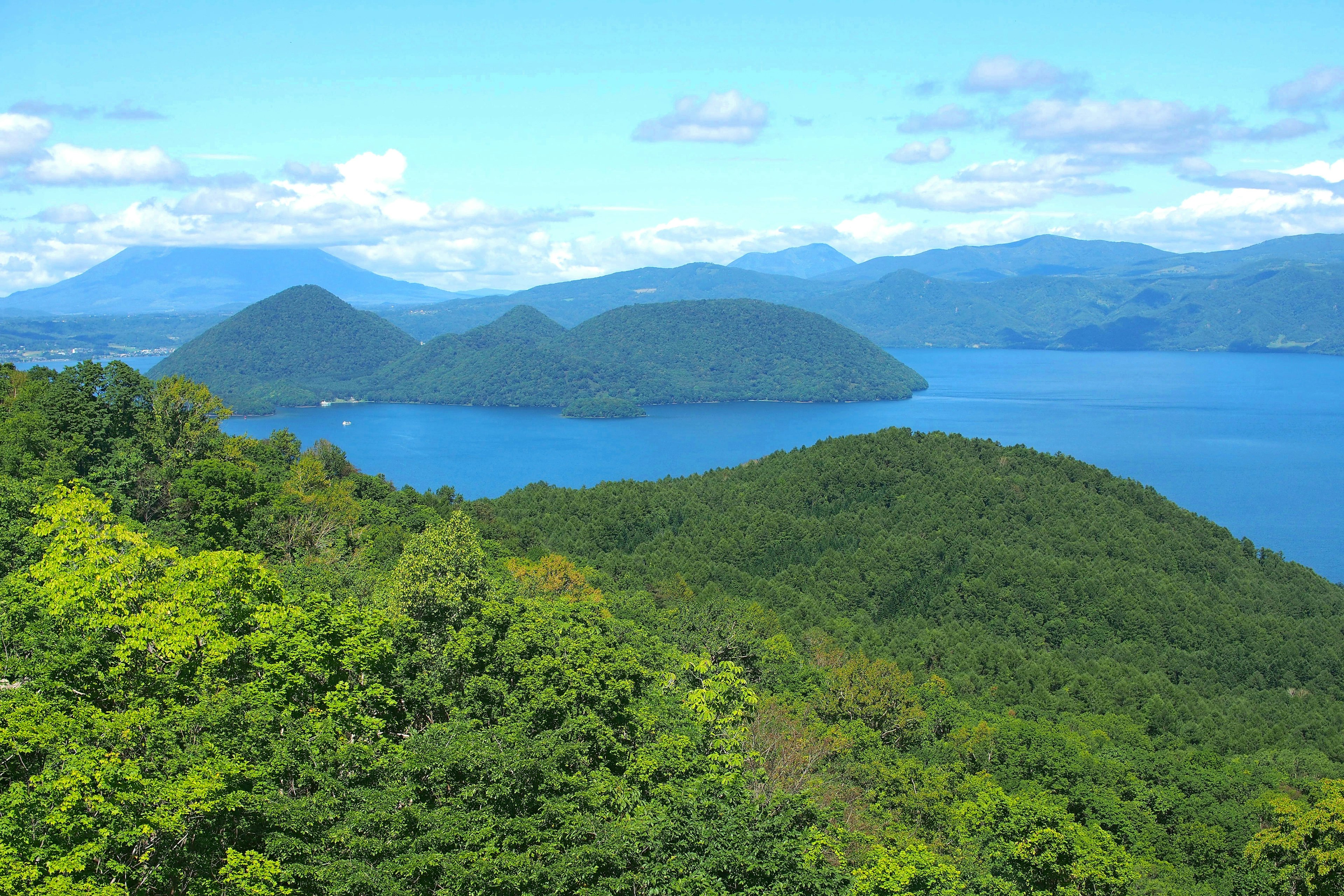 Malersiche Aussicht auf einen See umgeben von üppigen grünen Hügeln und Bergen unter einem klaren blauen Himmel