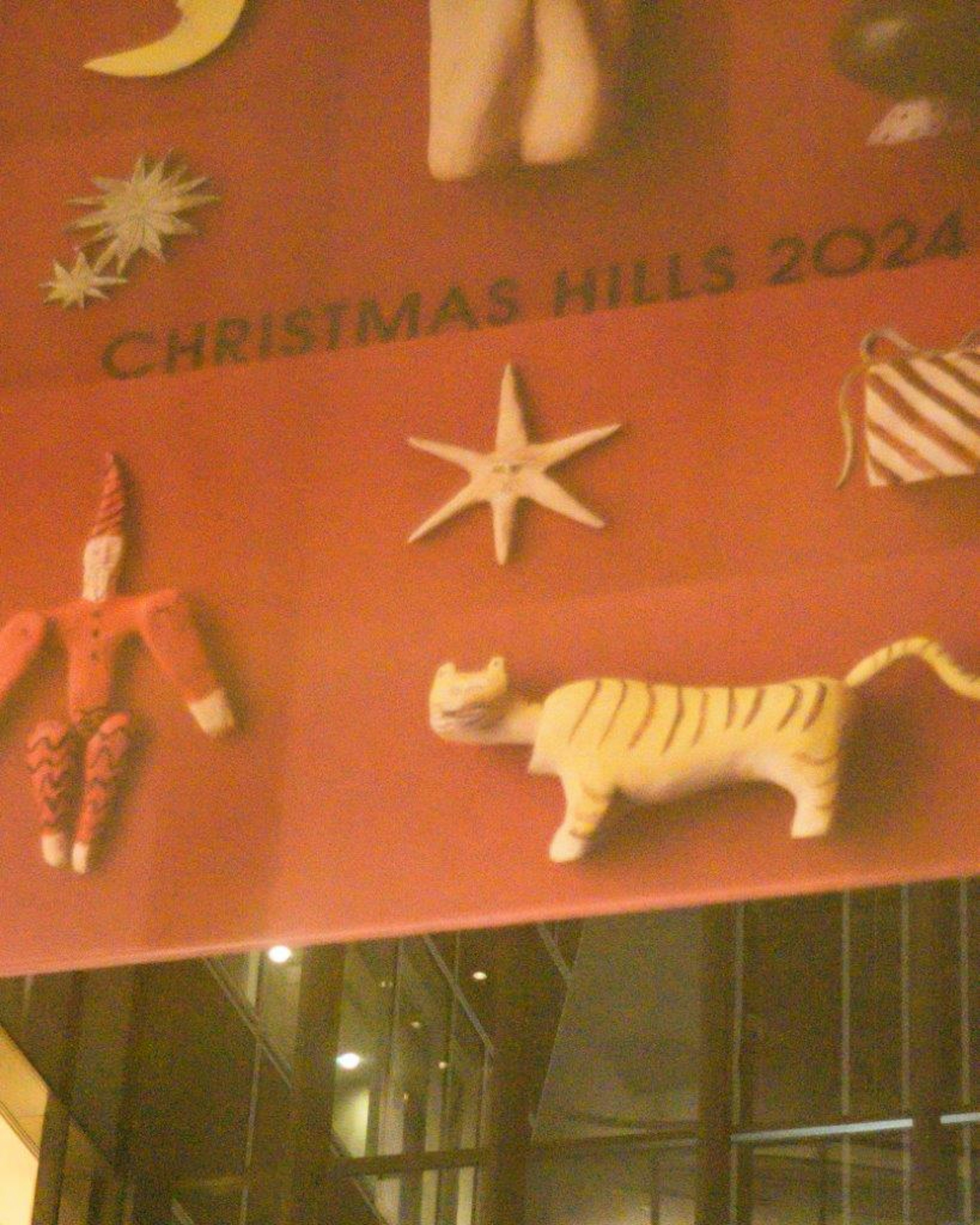 Barang dekoratif di latar merah dengan Christmas Hills 2024 termasuk bintang, kucing, dan figur Santa