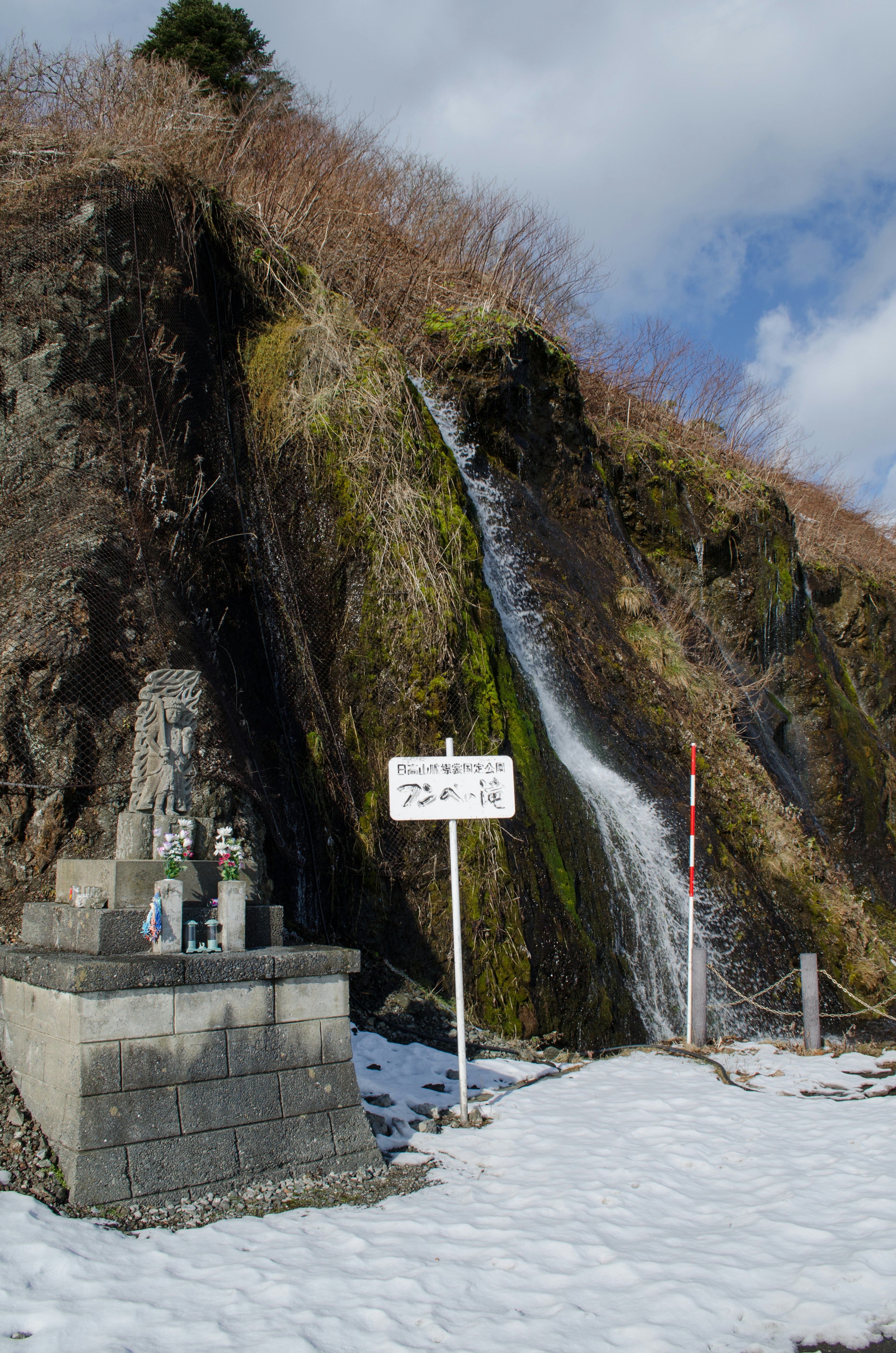 雪に覆われた岩肌と小さな滝のある風景 除雪された道と標識が見える