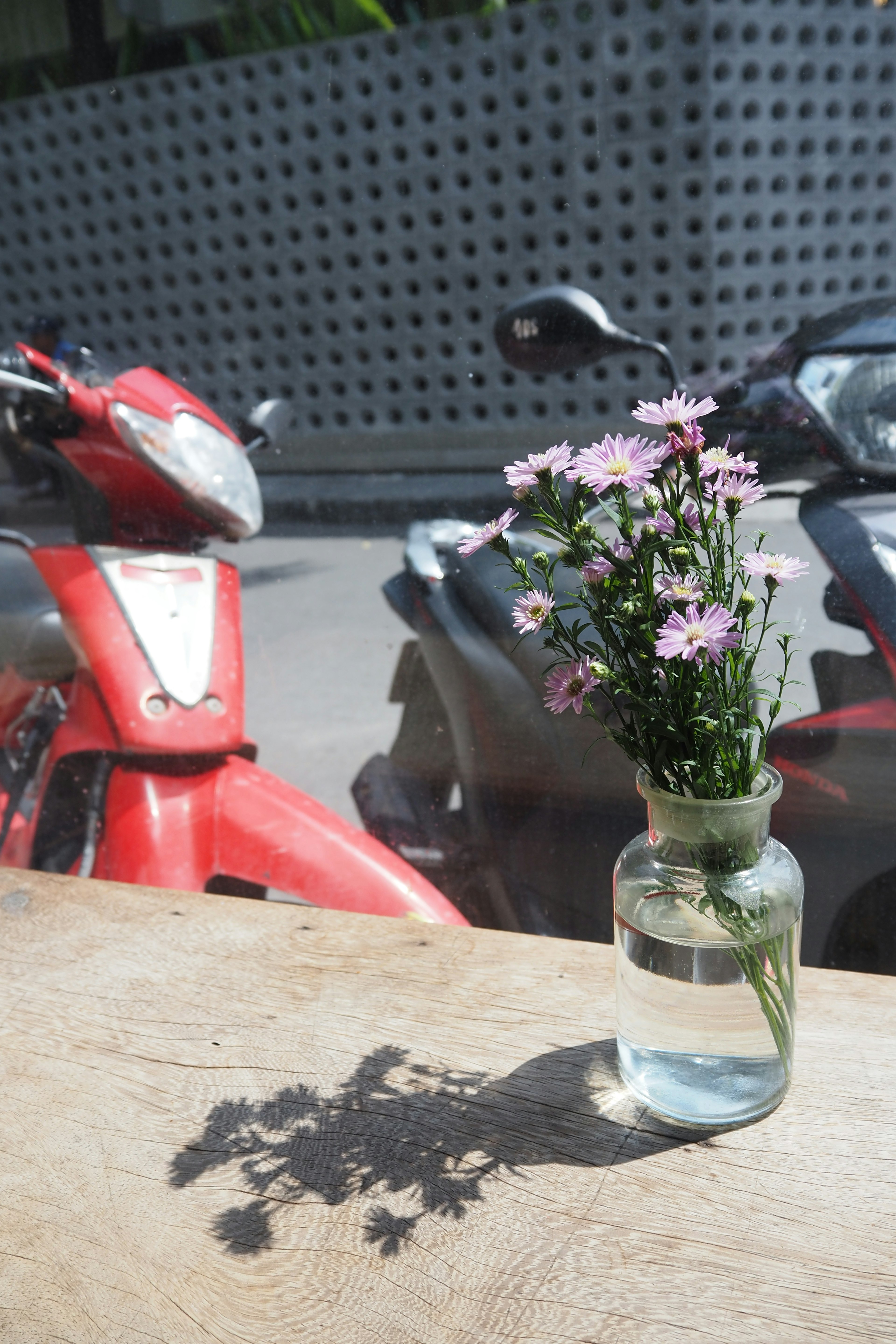 Small vase with tiny flowers casting a shadow with red motorcycle in the background