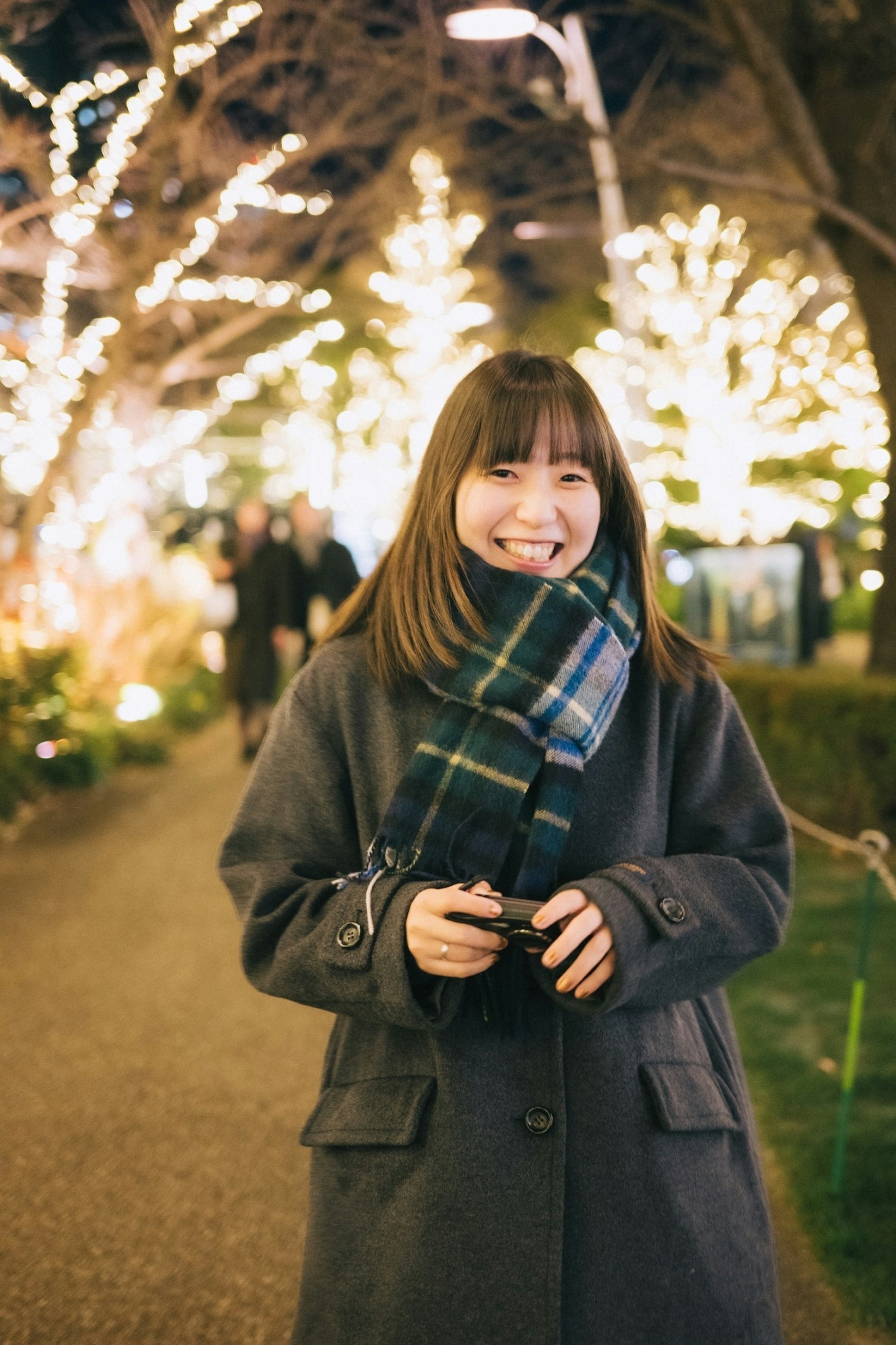 Donna sorridente in un cappotto caldo che tiene una macchina fotografica in un parco decorato con luci brillanti