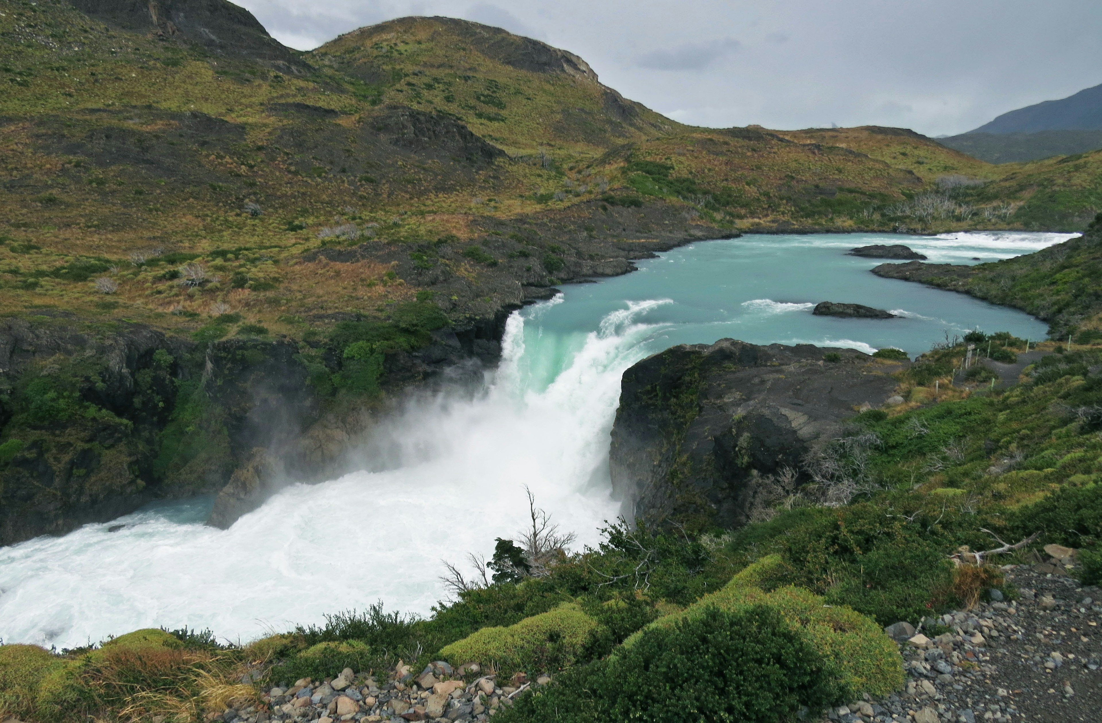 Cascata panoramica che si tuffa in acque turchesi circondata da verde lussureggiante e colline