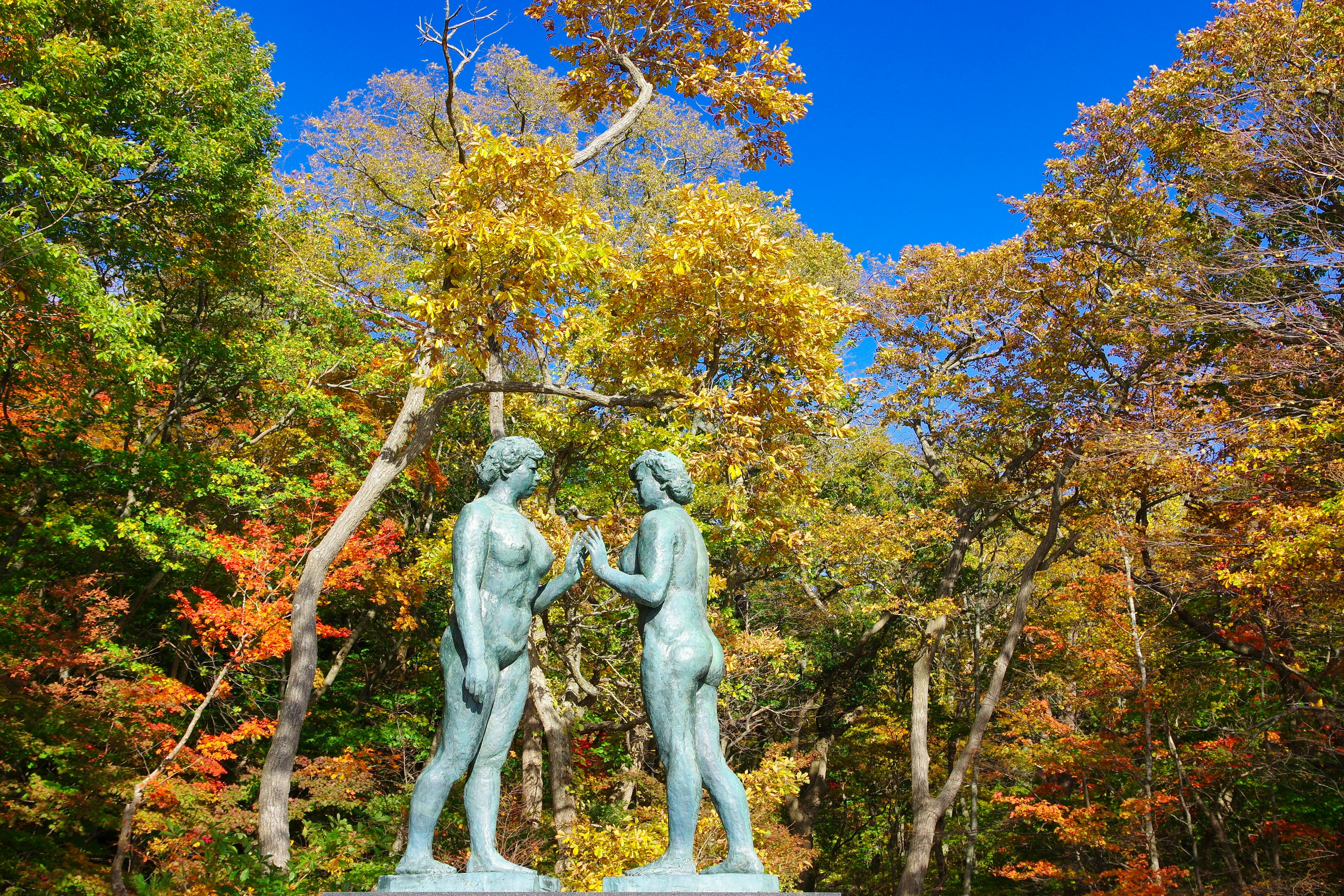 Zwei Statuen stehen unter einem blauen Himmel umgeben von herbstlichem Laub