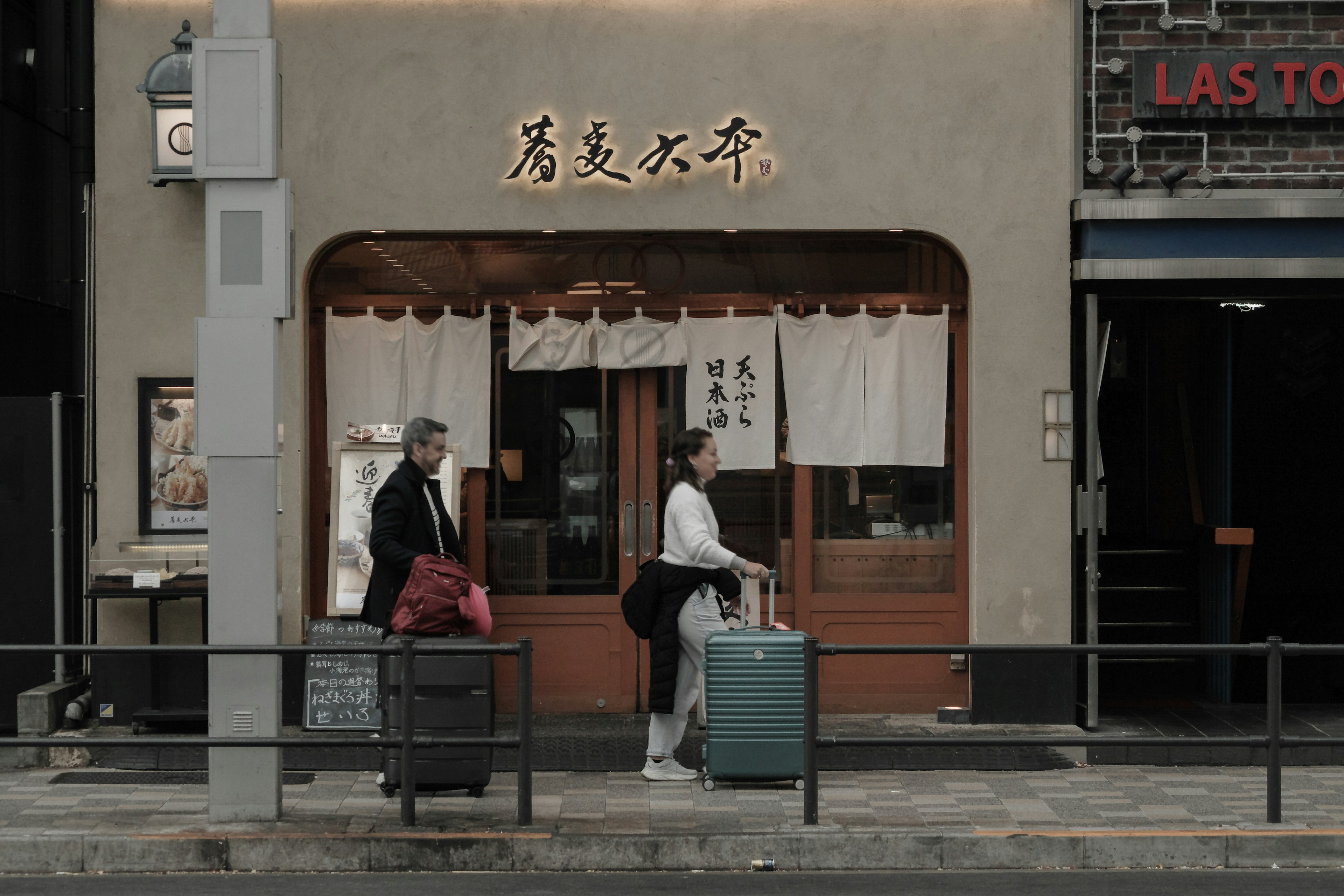 Dos personas con maletas de pie frente a una tienda japonesa tradicional en un entorno urbano