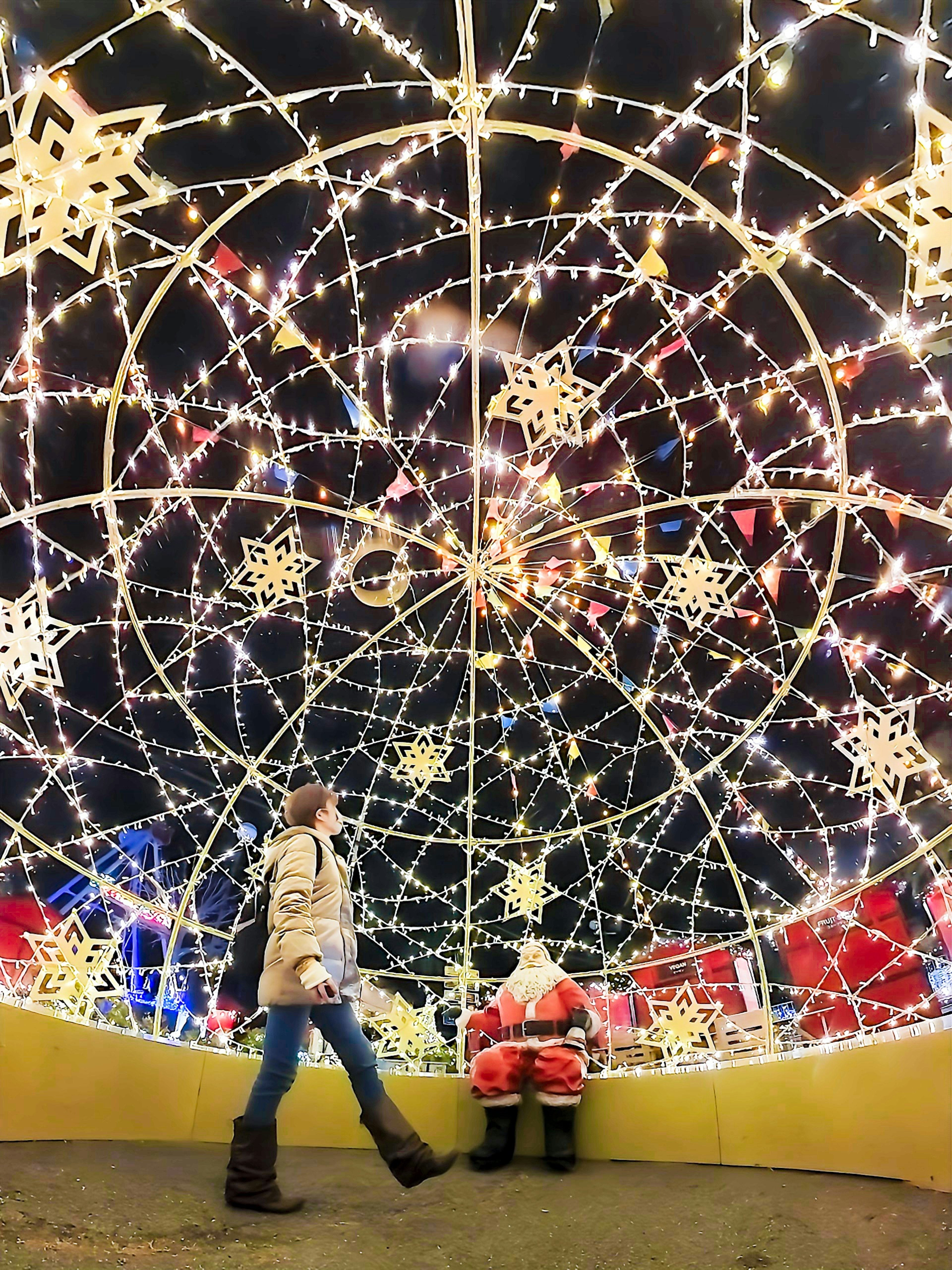 Una mujer caminando cerca de un niño sentado junto a Santa Claus rodeado de luces navideñas brillantes