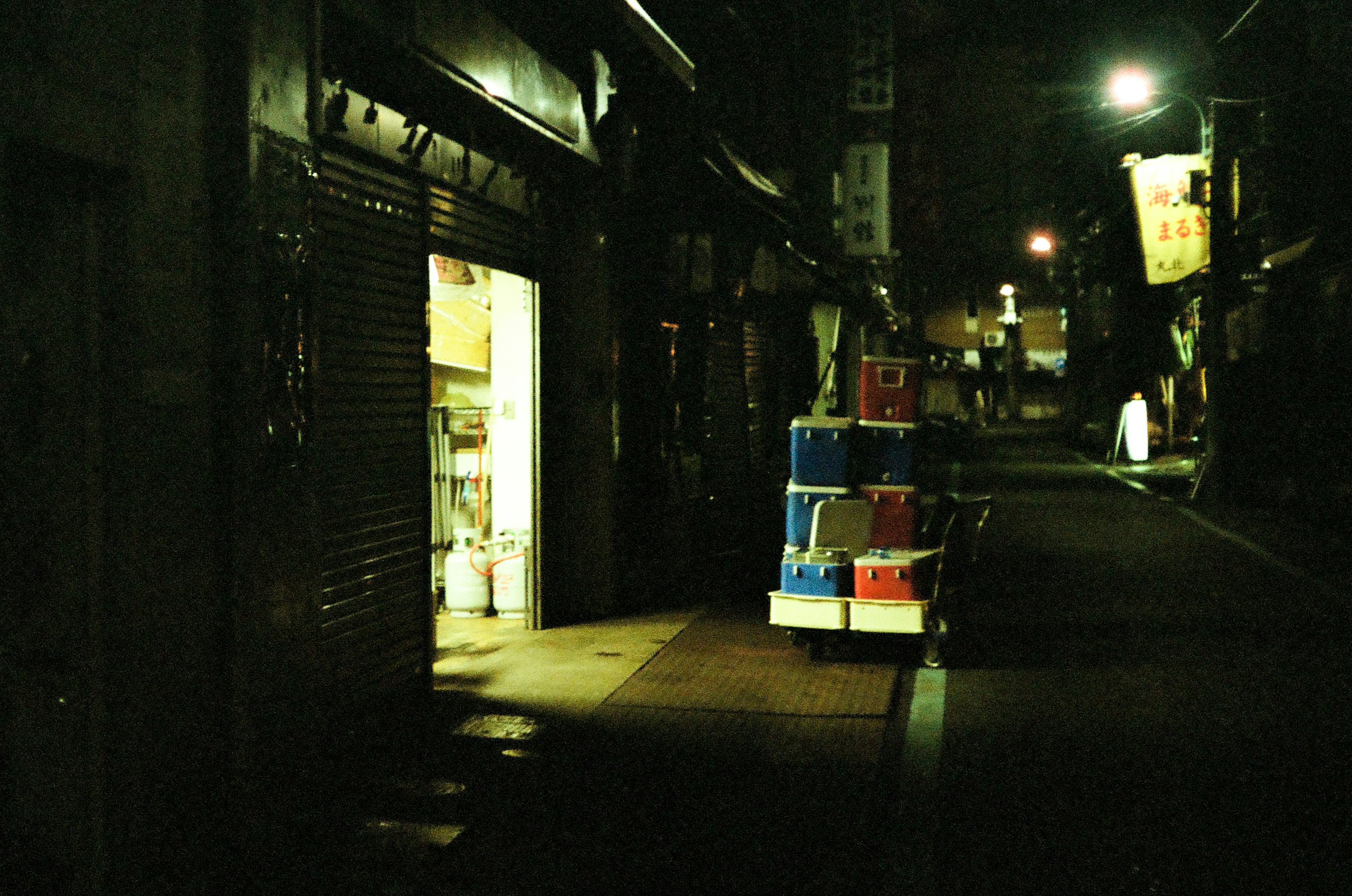 Dark street in a night market with a brightly lit store and stacked boxes