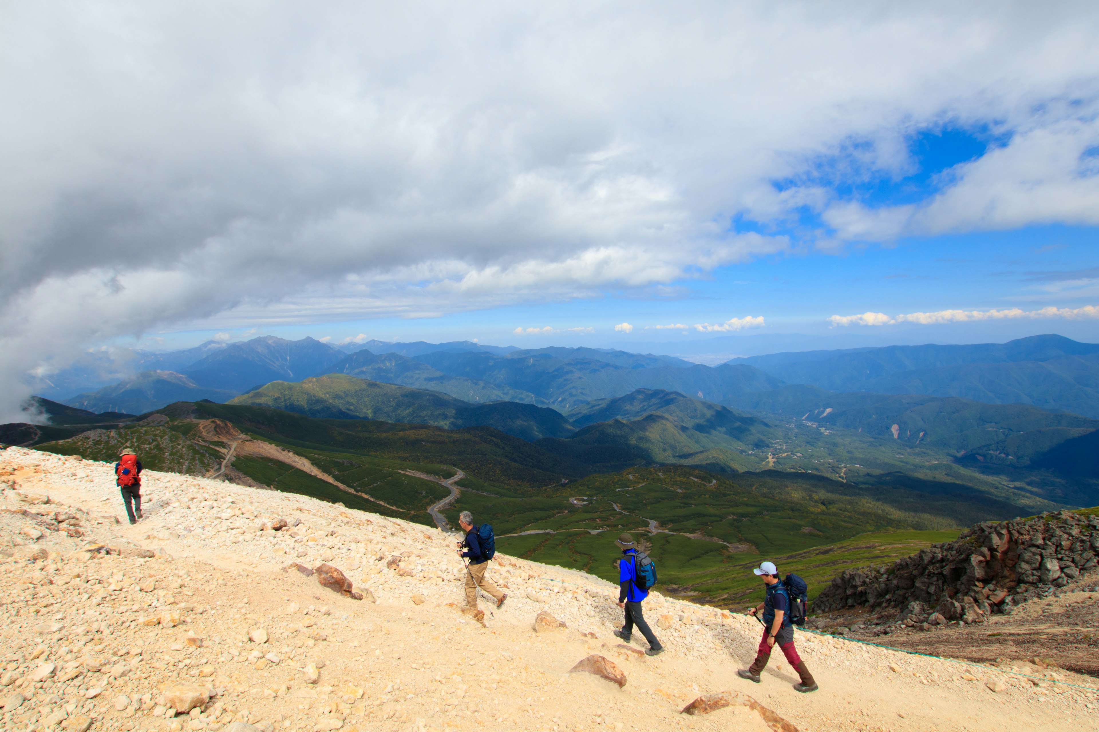 在藍天和雲朵下，登山者沿著山路行走 背景中有綠色的山丘