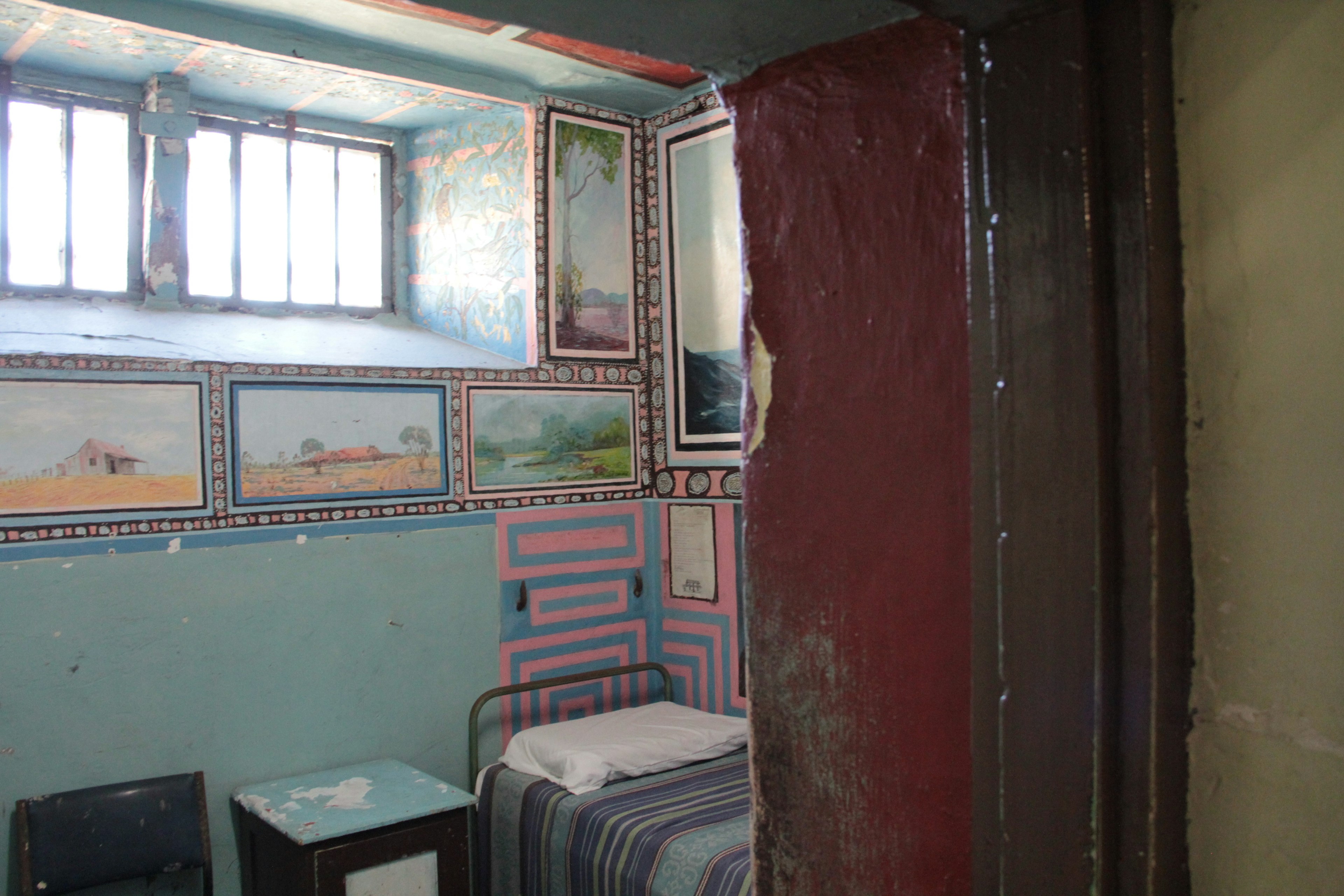 Interior de una pequeña habitación con paredes azules, pinturas, una cama individual y una mesa