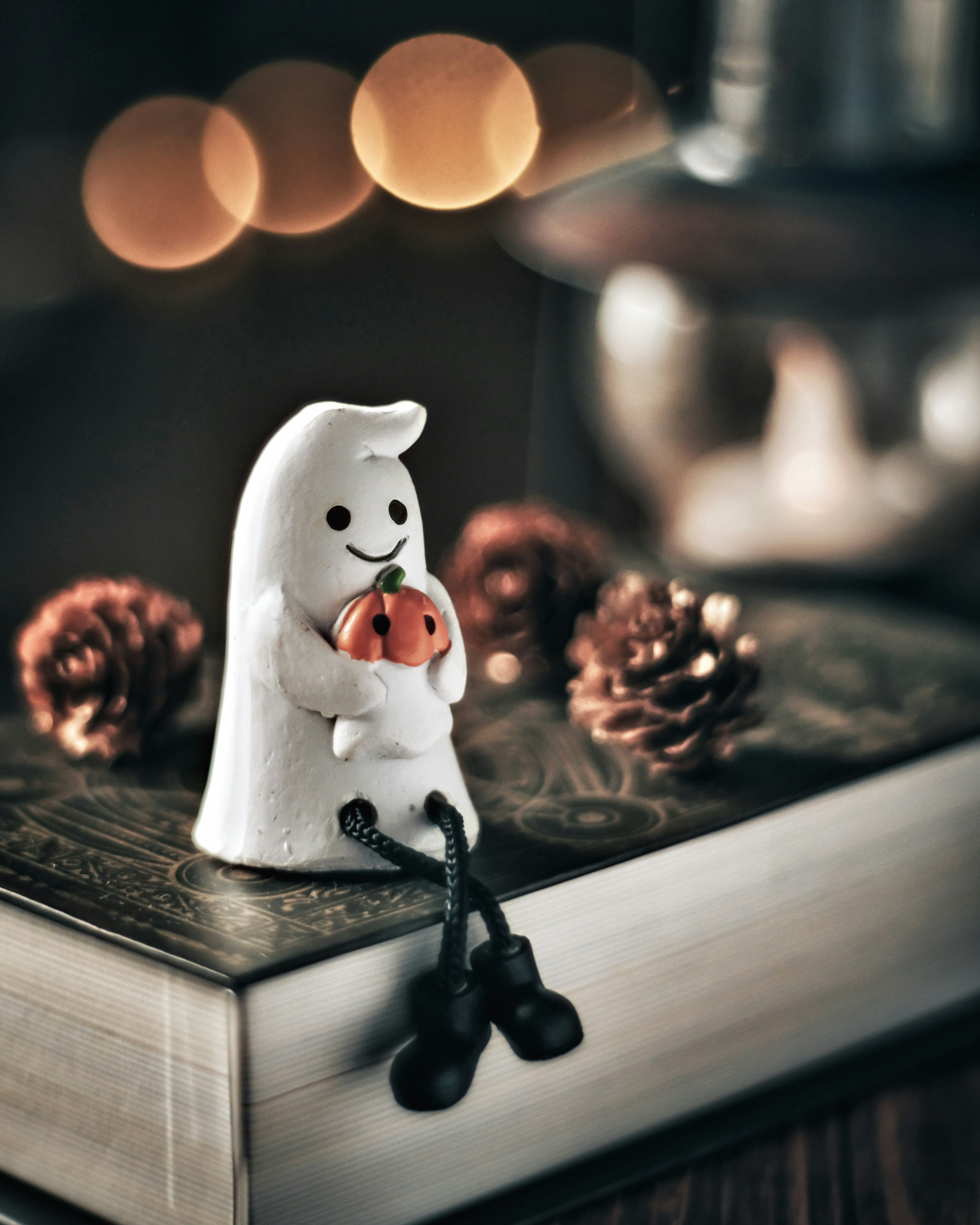 A white ghost figurine holding a small pumpkin The background features soft lights and pine cones