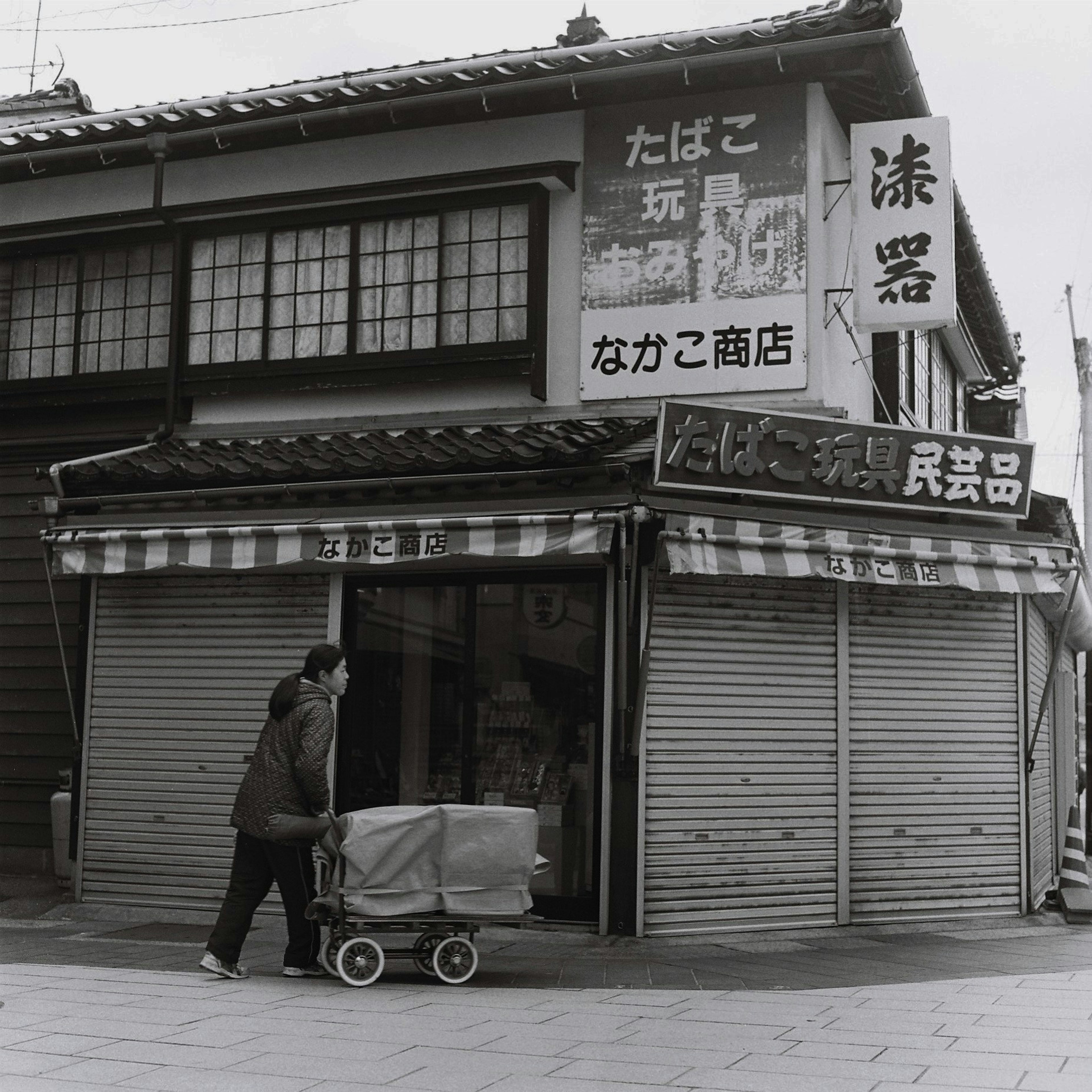 Foto en blanco y negro de una calle comercial con una mujer empujando un carrito y una tienda cerrada