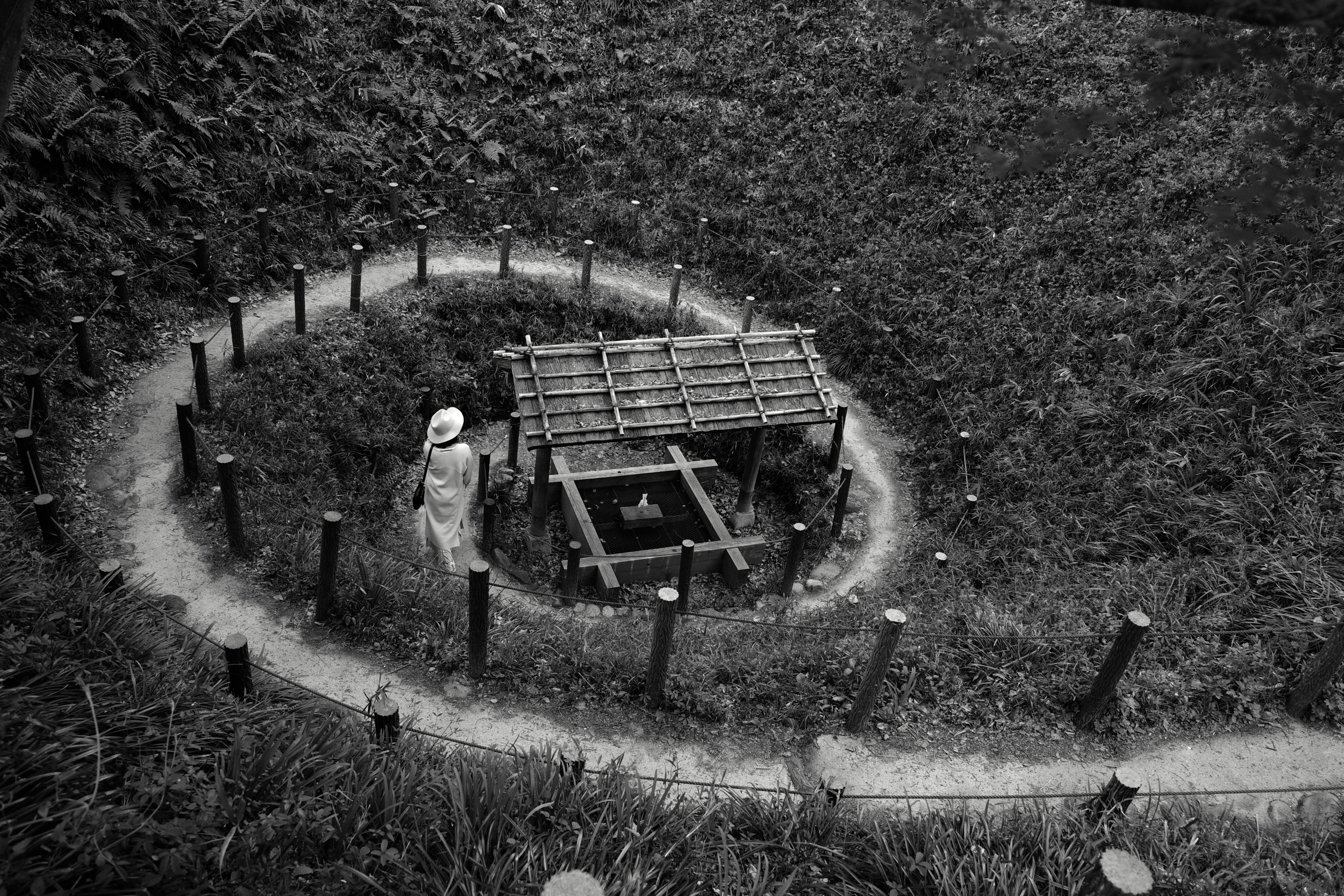 Eine Figur in der Nähe einer Bambusstruktur, umgeben von einem runden Weg in einer schwarz-weißen Landschaft