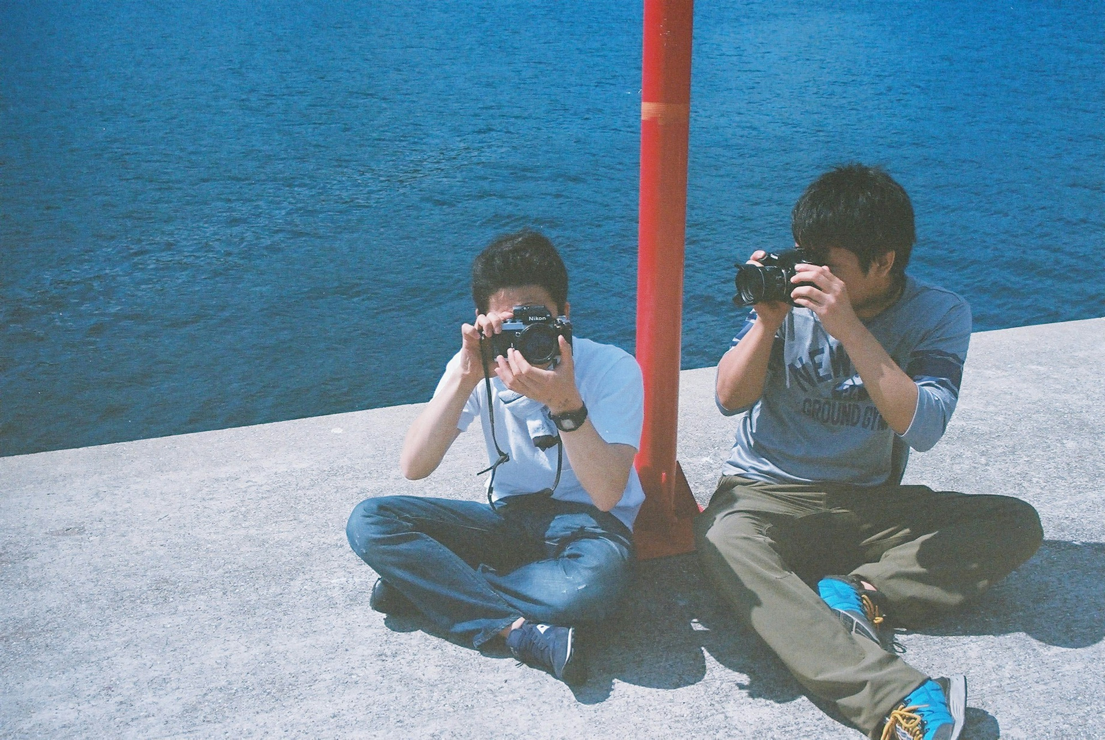 Two young people sitting on a concrete surface holding cameras with blue water and a red pole in the background