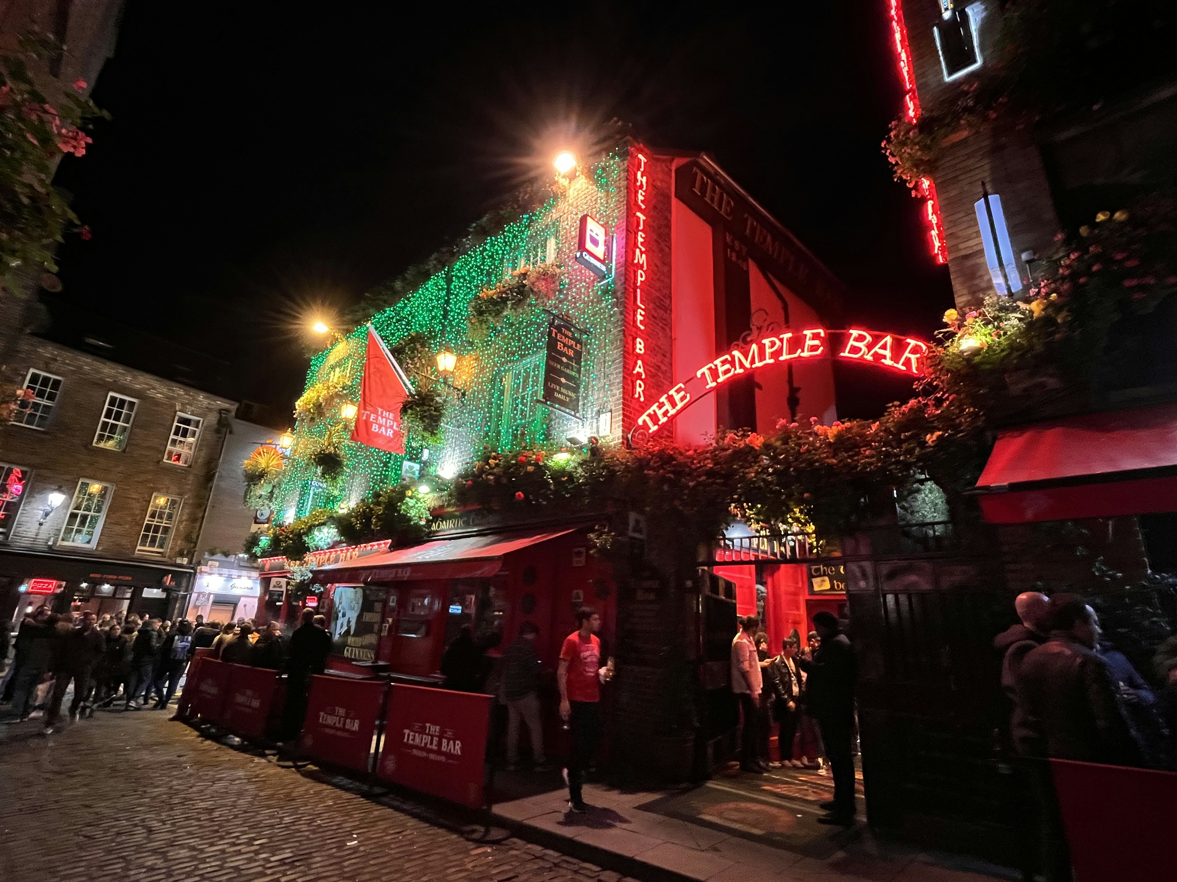 The Temple Bar exterior at night Bright neon sign and green decorations Crowds of people gathered