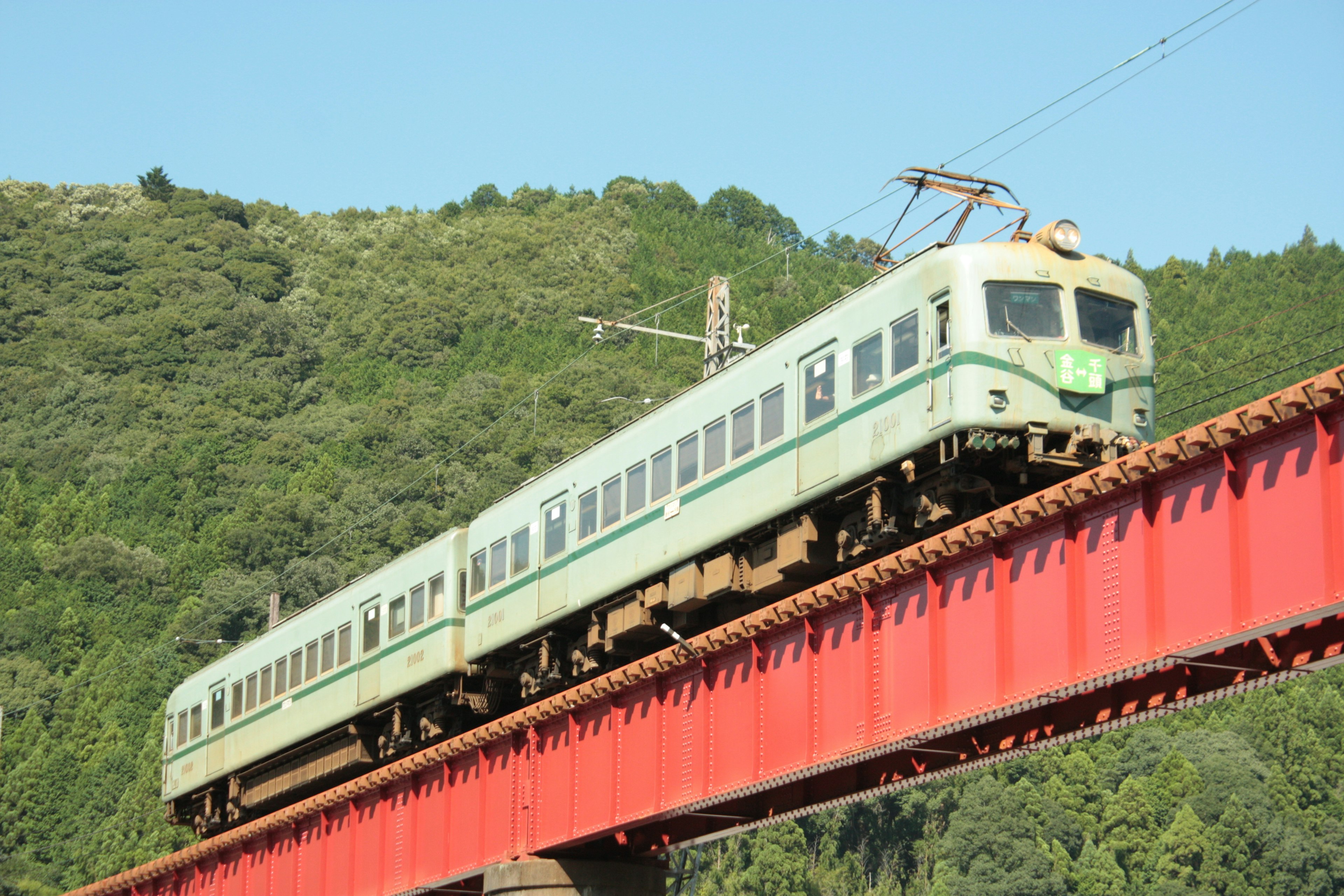 Sebuah kereta hijau melintasi jembatan merah dengan latar belakang gunung hijau