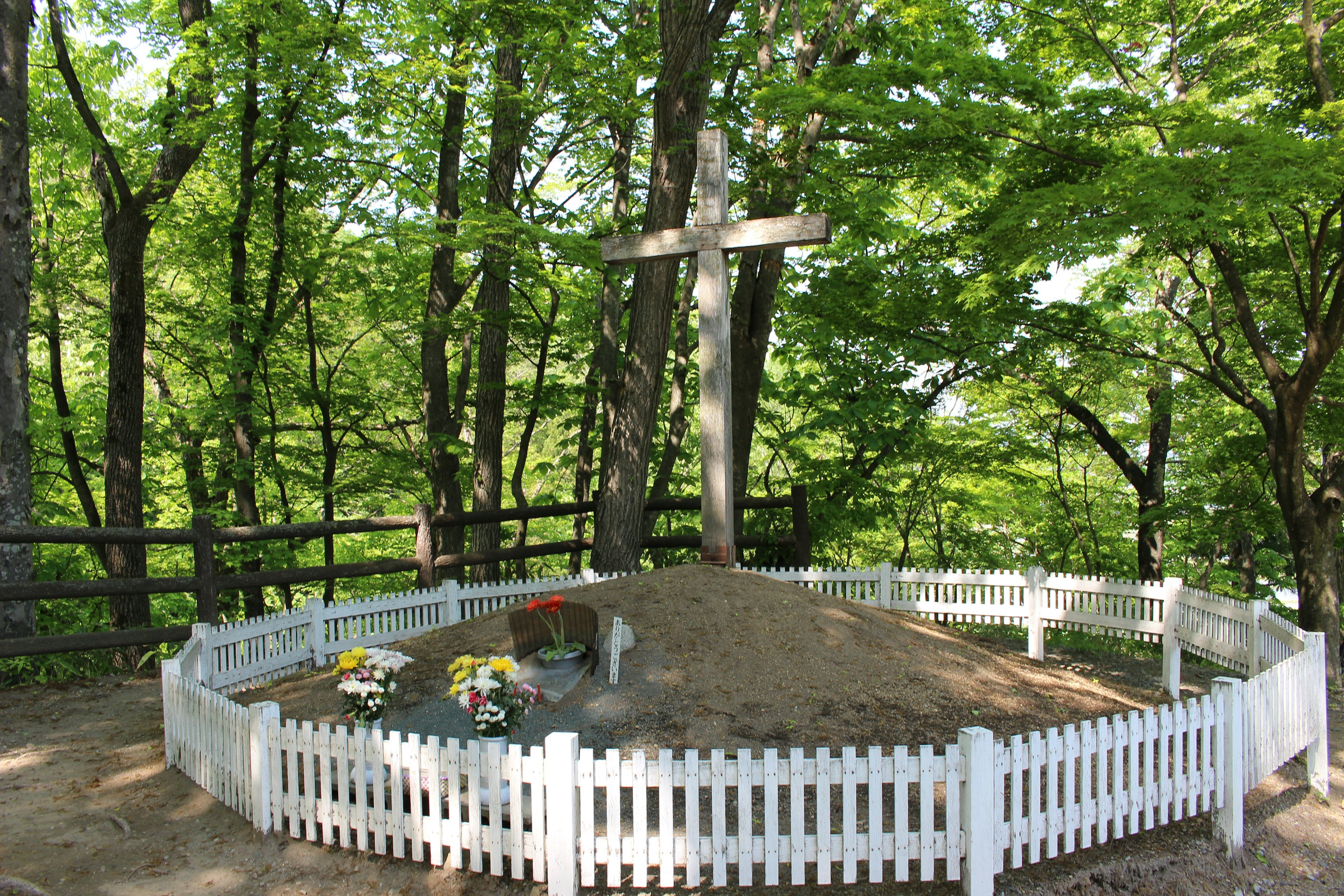 A small grave surrounded by lush green trees with a white fence and a cross