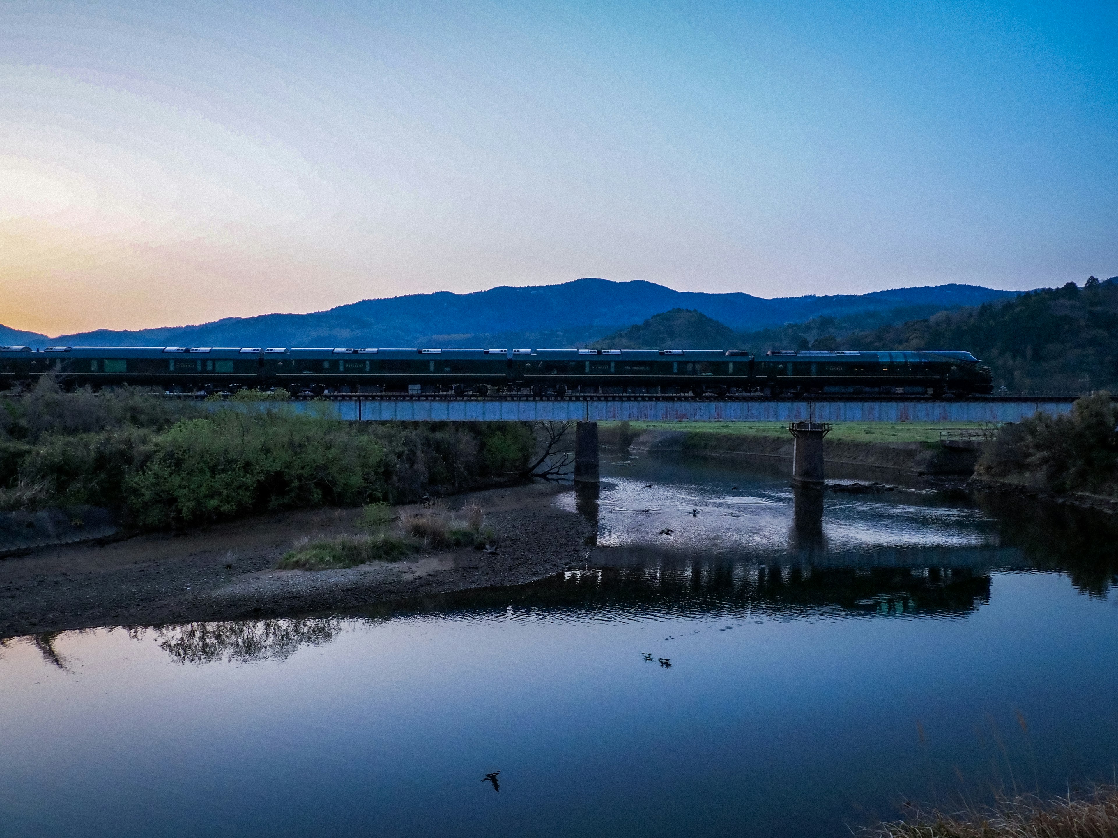 火車在日落時穿越河上的橋，背景是山脈