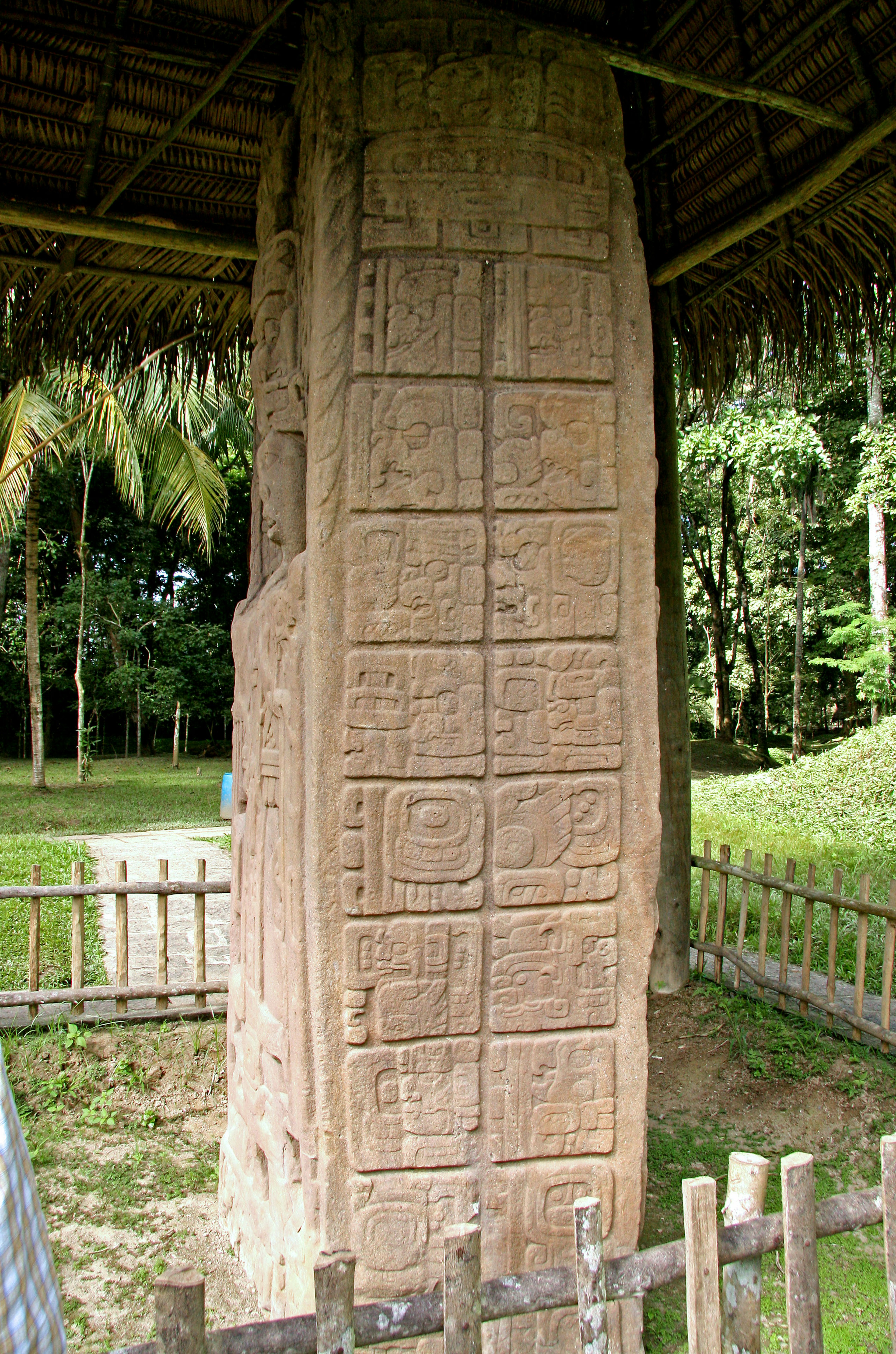 Alter Steinmonument mit Schnitzereien unter einem Strohdach umgeben von Grün