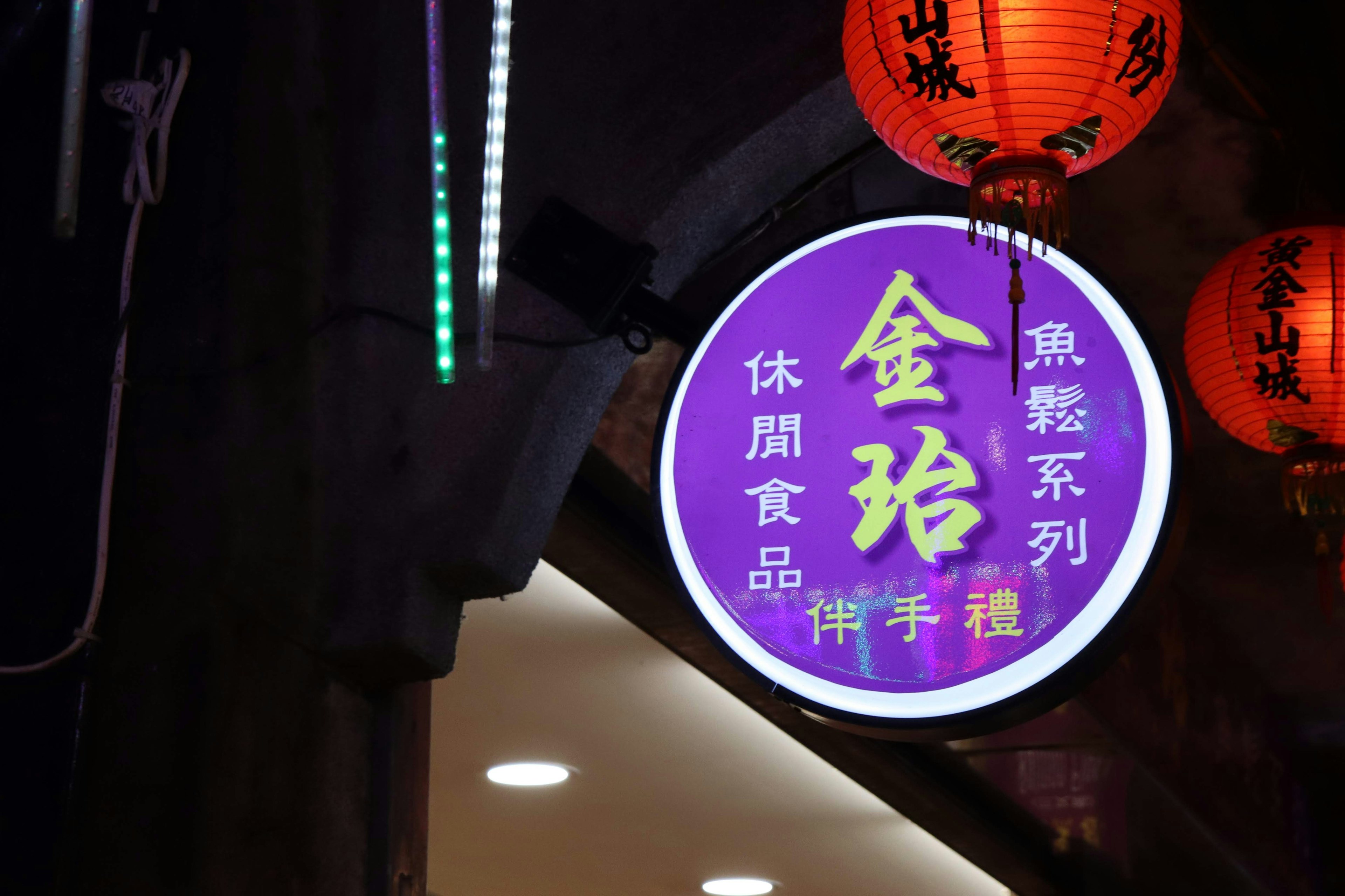 Escena nocturna con un letrero morado con letras doradas del nombre de una tienda rodeada de linternas rojas