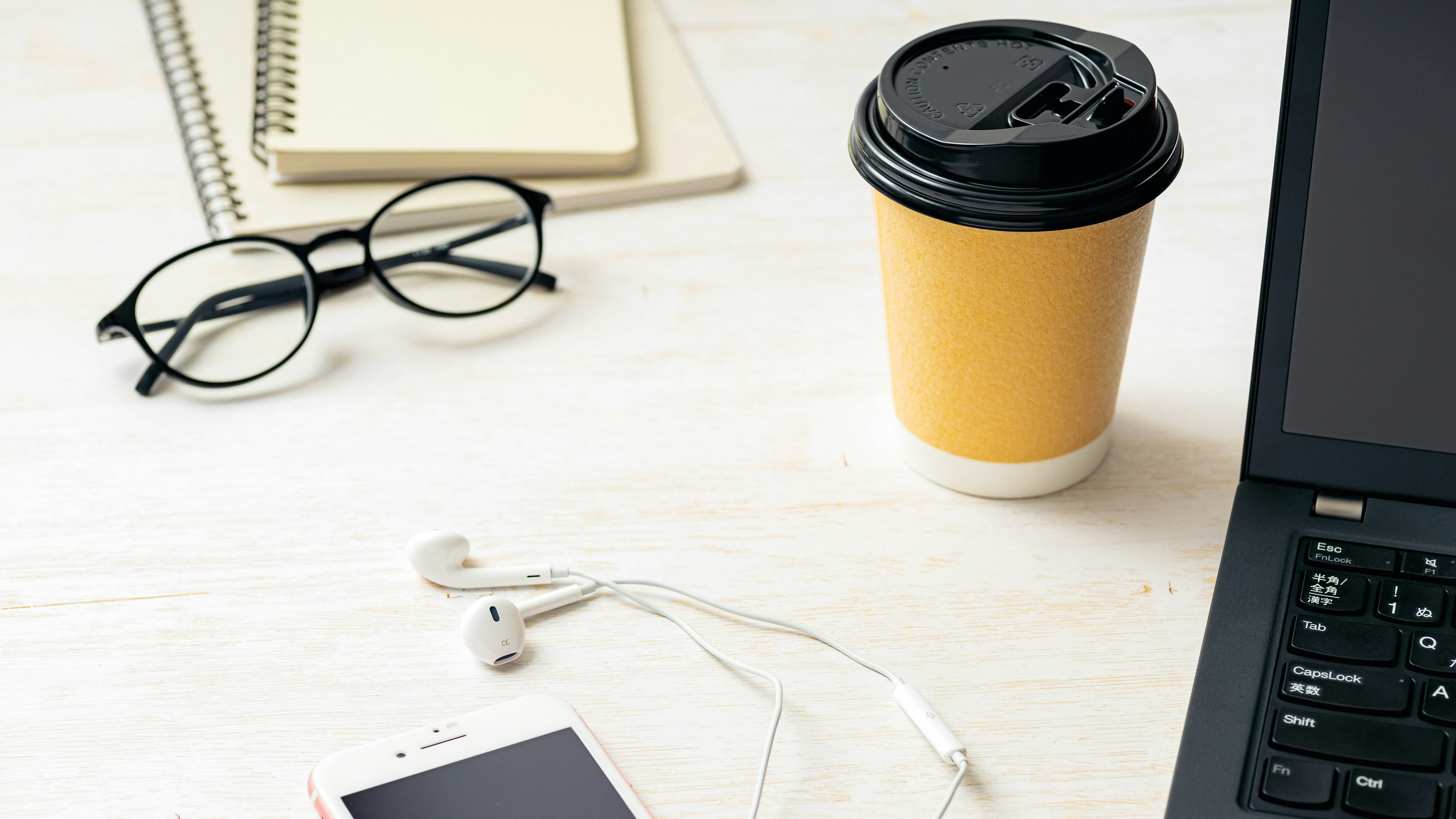 A laptop, coffee cup, notebook, glasses, earphones, and smartphone on a desk