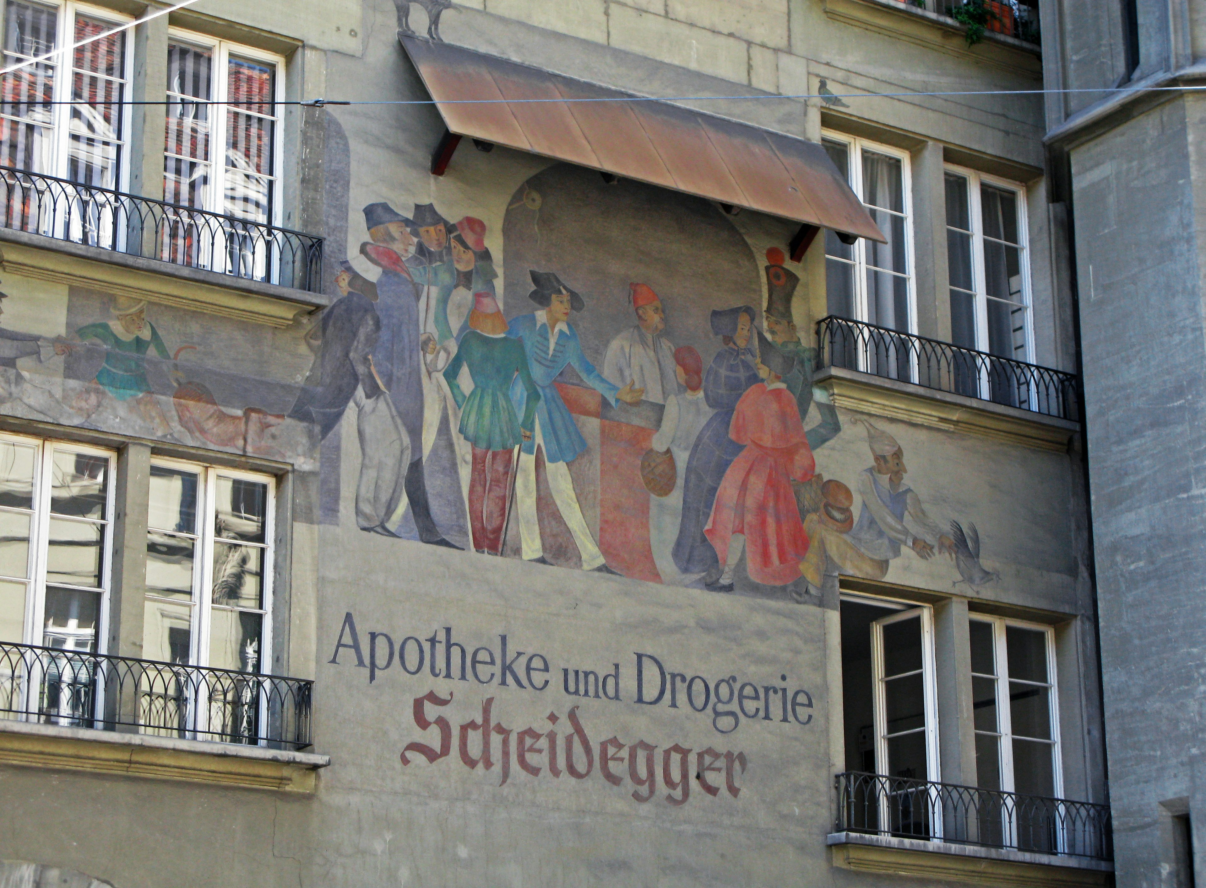 Fresco historique sur un mur de bâtiment en Suisse avec une enseigne de pharmacie et des gens rassemblés