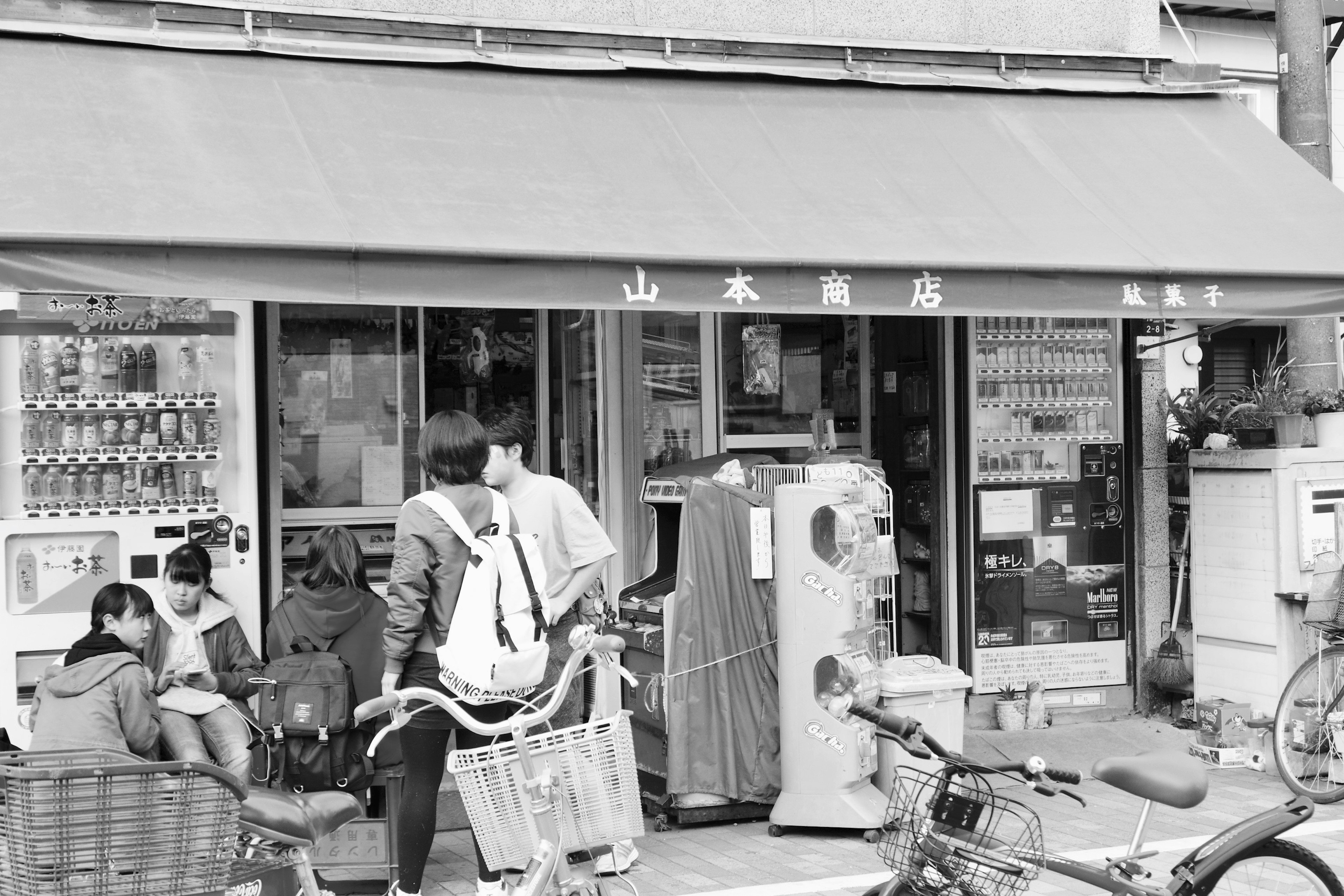 Black and white exterior of a small shop with several people around and bicycles parked nearby