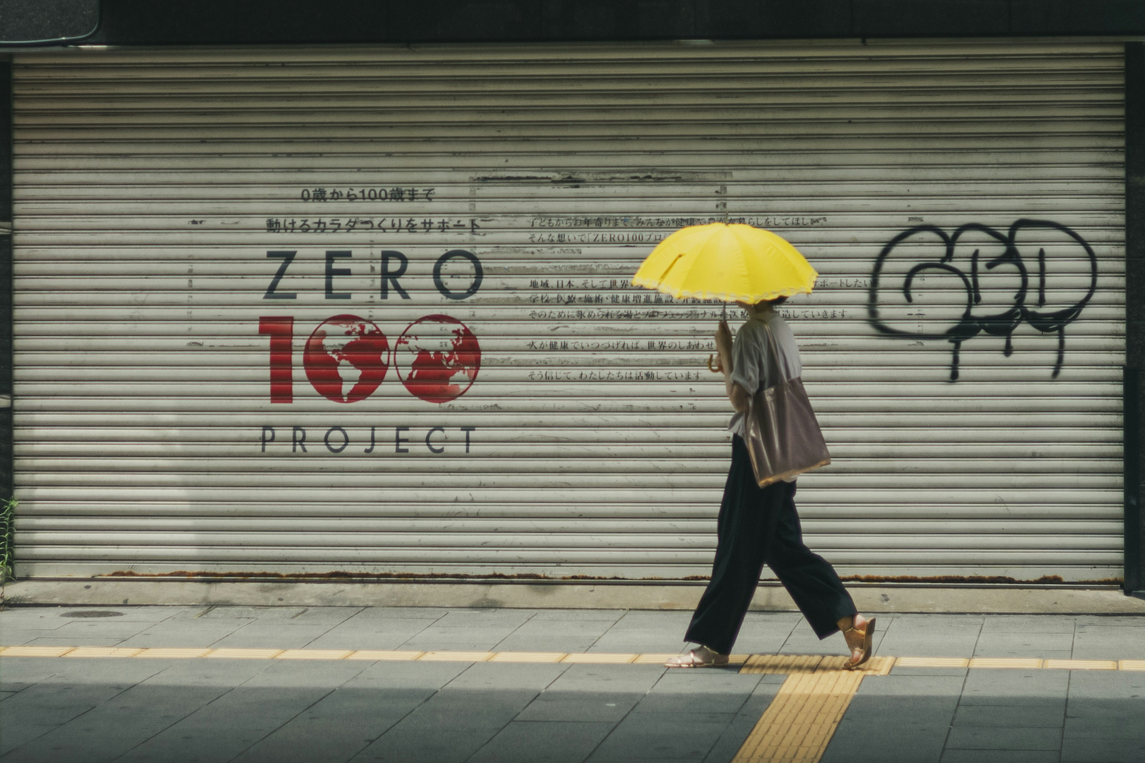 Woman walking with yellow umbrella in front of graffiti reading zero 100