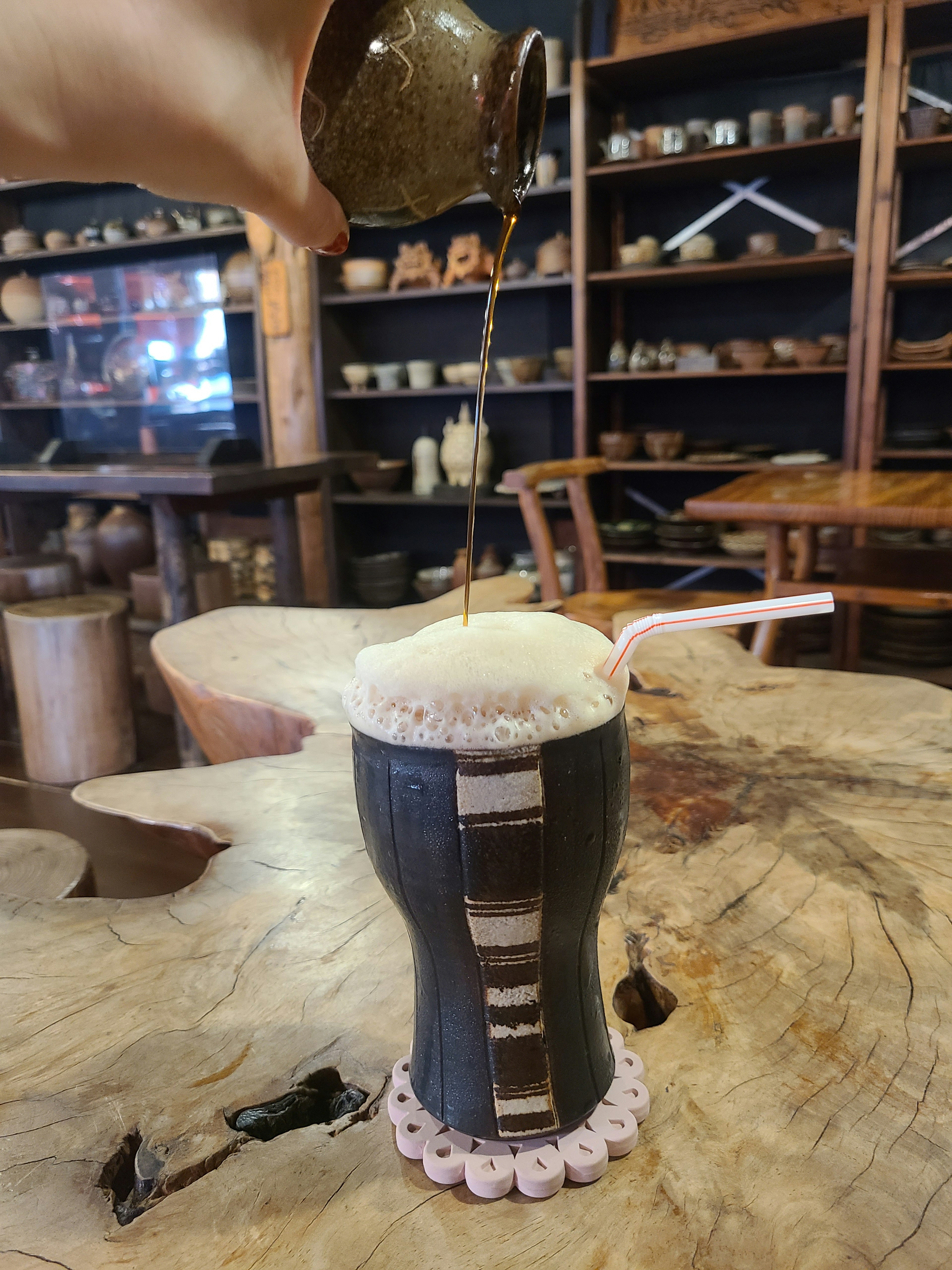 Una mano vertiendo una bebida en una taza de diseño único sobre una mesa de madera rodeada de cerámica al fondo