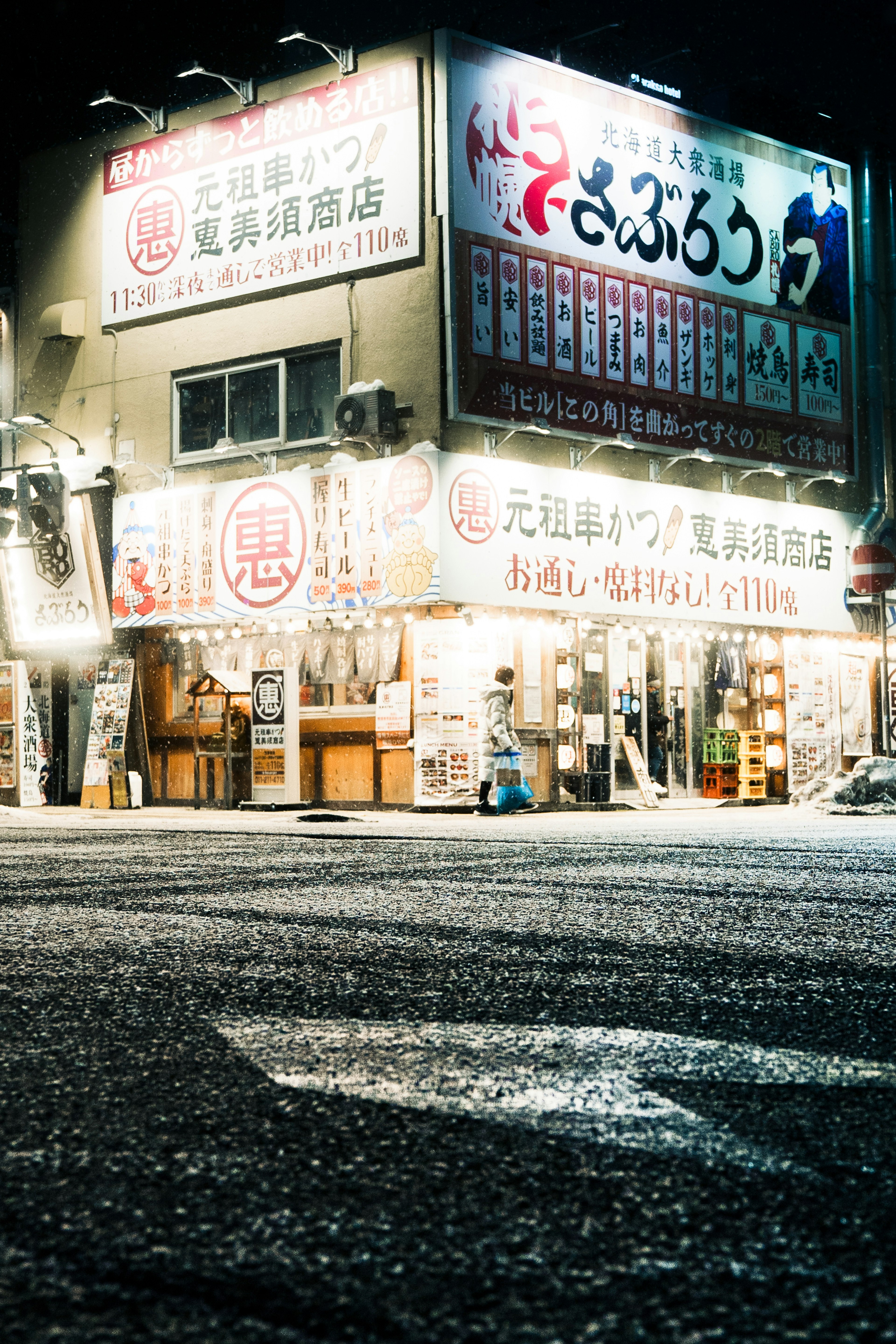 Eksterior izakaya di malam hari dengan papan nama dan tampilan menu yang terang